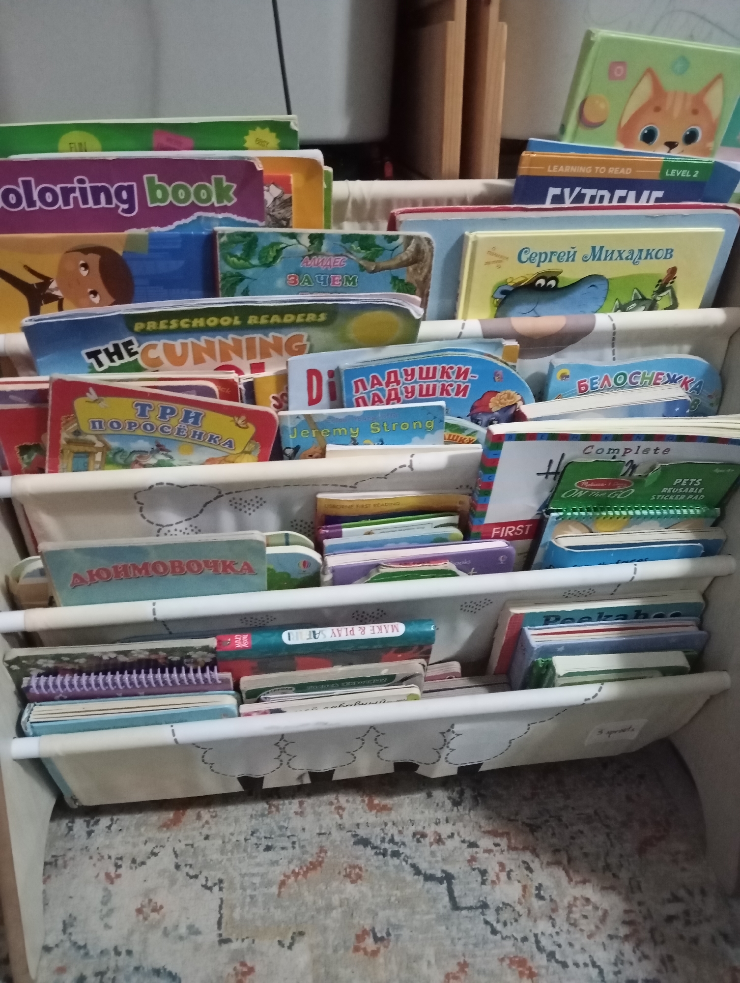 The image shows a close-up, eye-level, indoor shot of a white fabric bookshelf filled with children's books. The bookshelf is divided into several tiers. The books are colorful and varied in size and thickness, with some spines facing outwards and others stacked horizontally. Visible titles include "Coloring book," "PRESCHOOL READERS THE CUNNING," "ТРИ ПОРОСЁНКА," "Jeremy Strong," "Лягушка-квакушка," "БЕЛОСНЕЖКА," "Complete," "PETS REUSABLE STICKER PAD," and "PEEKABOO." Some of the books are open, revealing colorful illustrations. The overall environment appears to be a domestic setting, possibly a child's room or play area. There are no people or animals visible in the image. The background is blurred, showing hints of a wooden shelf unit and a patterned rug on the floor. There is no indication of the city of Dubai or the United Arab Emirates in this image.