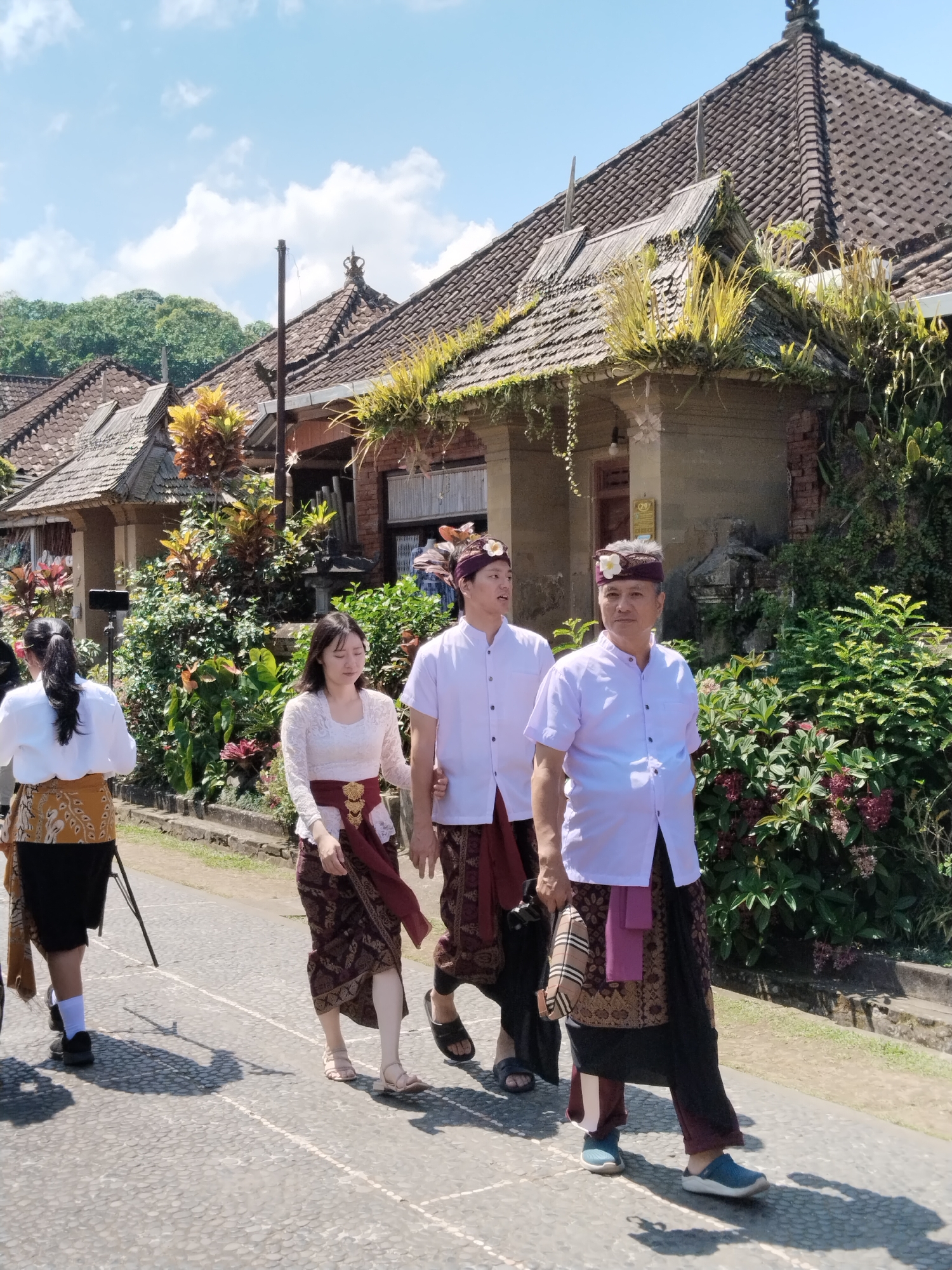 In the city of Bangli, Indonesia, three individuals are walking on a paved path. The two men in front are wearing white shirts and traditional Balinese sarongs. The man on the right has a purple head wrap adorned with a white flower, and the man to his left has a similar head wrap with a pink flower. They are walking alongside a woman who is wearing a white lace top and a dark red patterned sarong. To the left of this group, another person, likely a woman, is walking away from the camera, wearing a white shirt and a patterned sarong. In the background, traditional Balinese houses with tiled roofs and overgrown vegetation are visible. The sky is blue with scattered clouds.