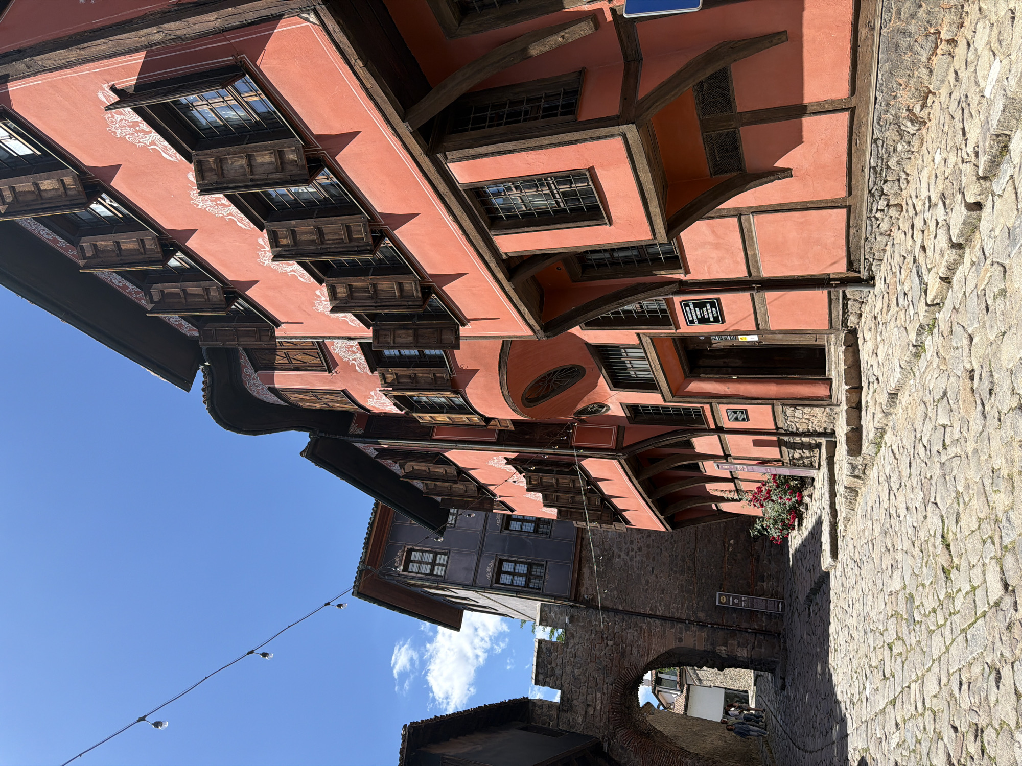The image captures a narrow street in the historic Old Town of Plovdiv, Bulgaria. The buildings are predominantly half-timbered with salmon-colored plaster. The buildings have wooden window frames and projecting windows on the first few floors. A stone wall and archway partially obscure a building in the background. Above the wall, a blue building stands out. A string of lights stretches across the sky on the left. The sky is blue with a few clouds. The ground is cobblestone. A sign is visible on one of the buildings. Flowers grow in a box under the buildings. People sit near the archway.