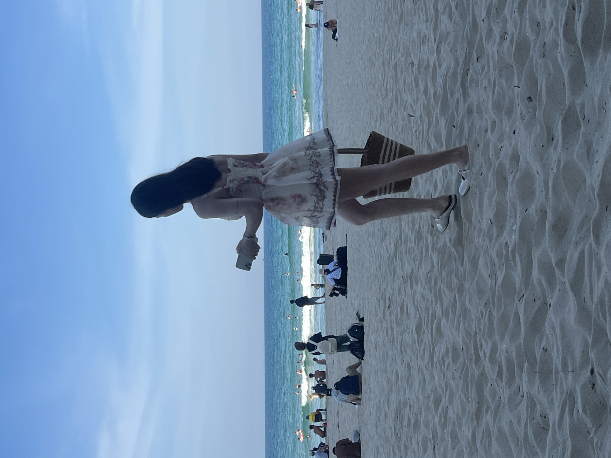 This media file depicts a vibrant beach scene in Đà Nẵng, Vietnam. The primary subject is a young woman with long, dark hair, wearing a short, floral-patterned dress and white sandals. She is standing on the sandy beach, holding a mobile phone in her right hand. Beside her, a straw beach bag with striped detailing is propped up.
The background reveals a picturesque coastal setting. The turquoise ocean, with gentle waves breaking on the shore, stretches out towards the horizon under a clear blue sky. Scattered across the beach and in the water are numerous other people enjoying the day. Some are swimming or wading in the sea, while others are relaxing or walking on the sand. The beach itself is covered in fine, light-colored sand, with ripples and undulations creating a textured surface.
The overall impression is one of a leisurely summer day, likely in the afternoon given the bright sunlight and clear sky. The mood is relaxed and cheerful, indicative of a typical beach outing. No text is visible in the image.