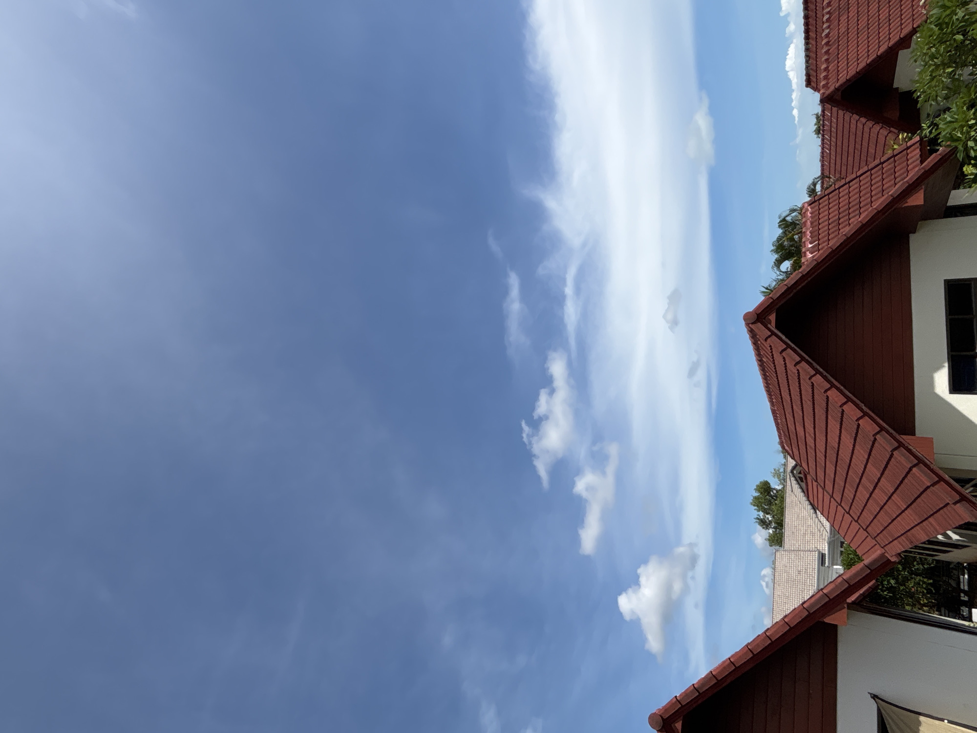 The image displays a row of red-tiled roofs against a bright blue sky with scattered white clouds. The roofs are angled, and portions of white-painted walls and brown siding are visible. Lush green foliage peeks out from between the buildings. The scene suggests a residential or commercial area with a clear, sunny day. The composition is dominated by the sky and the architectural elements of the rooftops.