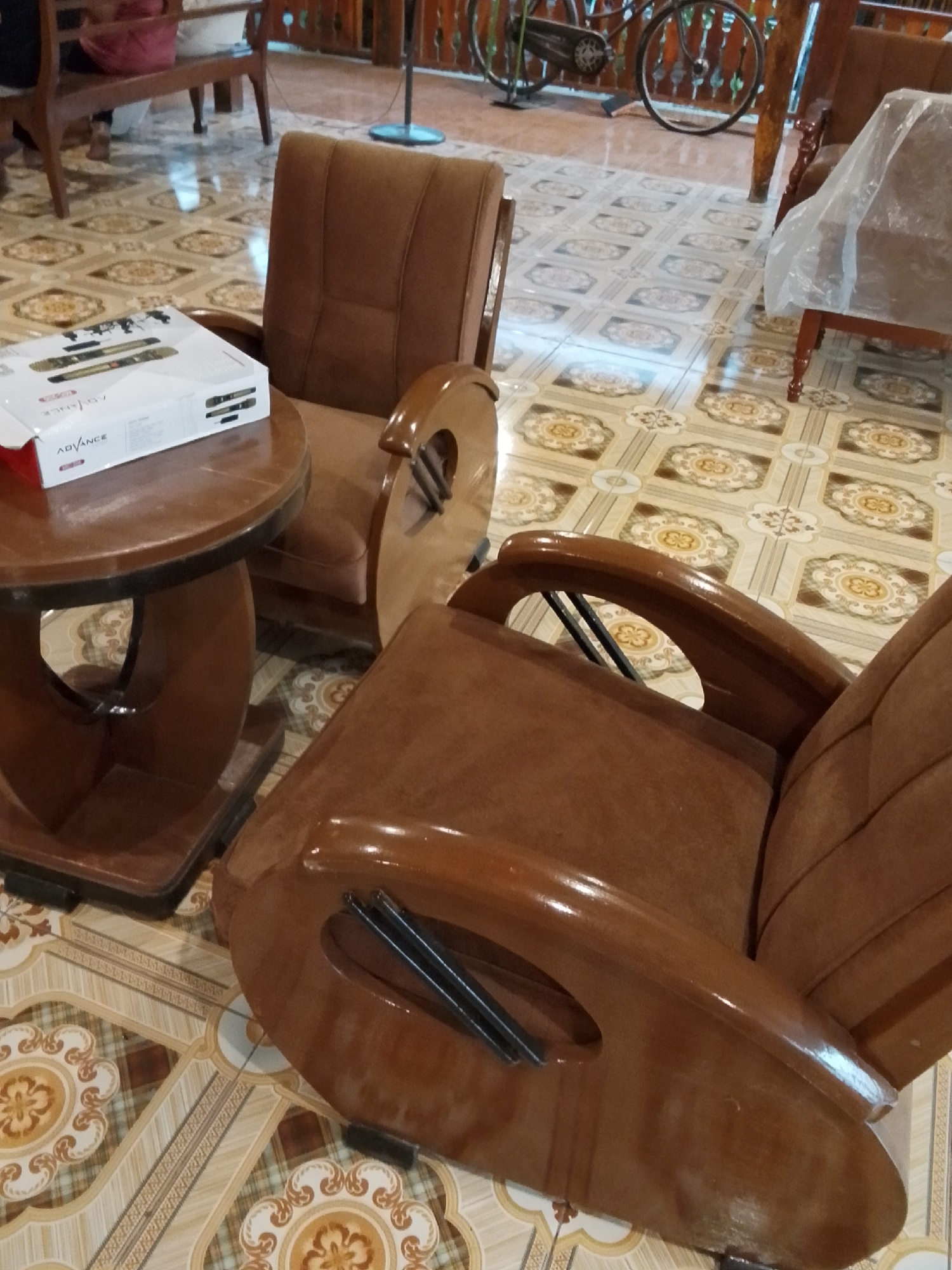 The image depicts an indoor scene with two brown upholstered chairs and a small round wooden table in the foreground. On the table, a white box with the brand name "ADVANCE" is visible, containing two microphones. The chairs are made of wood with upholstered seats and backs. The flooring is tiled with a patterned design. In the background, a wooden bench with a red cushion is partially visible, as well as a bicycle and a standing fan. The setting appears to be a residential or public space in Mojotengah, Indonesia, possibly a waiting area or a lounge. The lighting suggests it is daytime. There are no people visibly interacting in the scene. The overall impression is one of a quiet, somewhat dated interior.