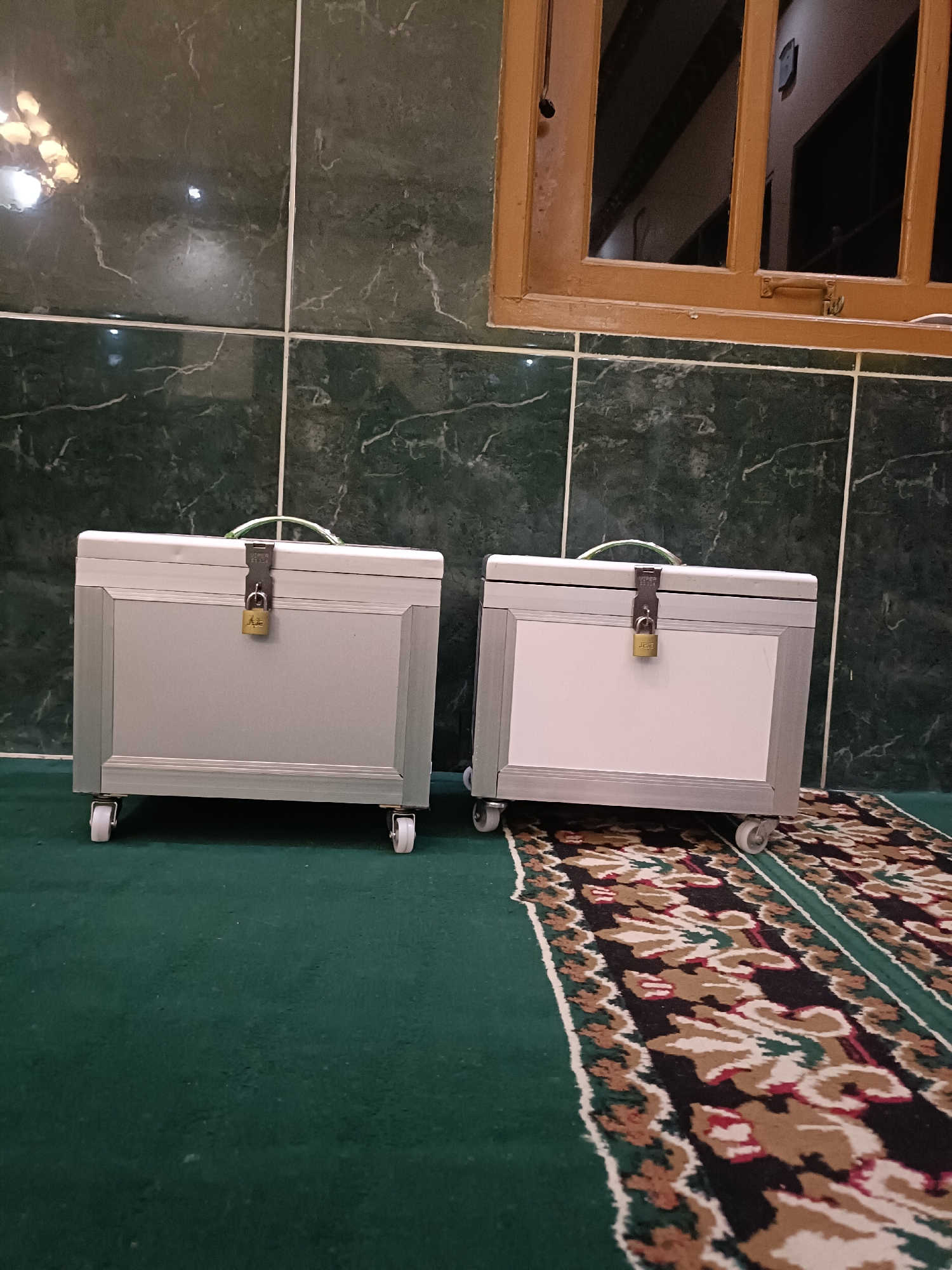 The image depicts an indoor scene, likely a room with tiled walls and a green carpet. Two identical, silver-colored storage trunks with white fronts are positioned on the carpet. Each trunk has a handle on top and a small padlock securing the front. Both trunks are equipped with four white casters, suggesting they are designed for easy mobility. The trunk on the left has a small, gold-colored padlock, while the one on the right has a similar padlock. A patterned carpet runs across the floor in front of the trunks, featuring a geometric design with colors like black, white, red, and brown. In the background, there's a window with a wooden frame and bars. The walls appear to be dark green tiles with white veining. The scene is lit by overhead lighting, which creates reflections on the tiled walls and the surfaces of the trunks. There are no people visible in the image. The location is identified as Kediri City, Indonesia.