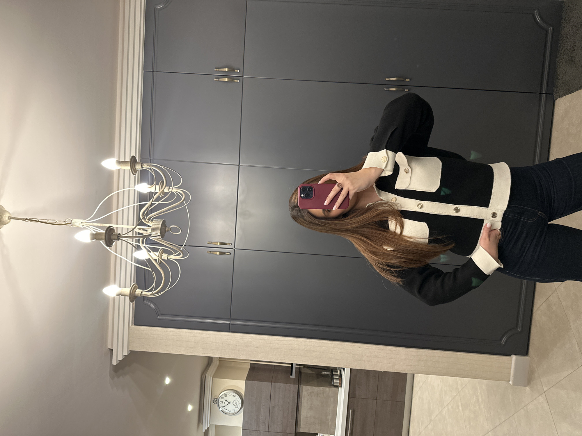 A person takes a selfie with a red phone in front of a dark grey cabinet. The person has long brown hair and is wearing a black and white jacket. A white chandelier is visible on the left side of the image. The background shows a kitchen with a clock on the wall. The location could be a house or apartment in Yerevan, Armenia.