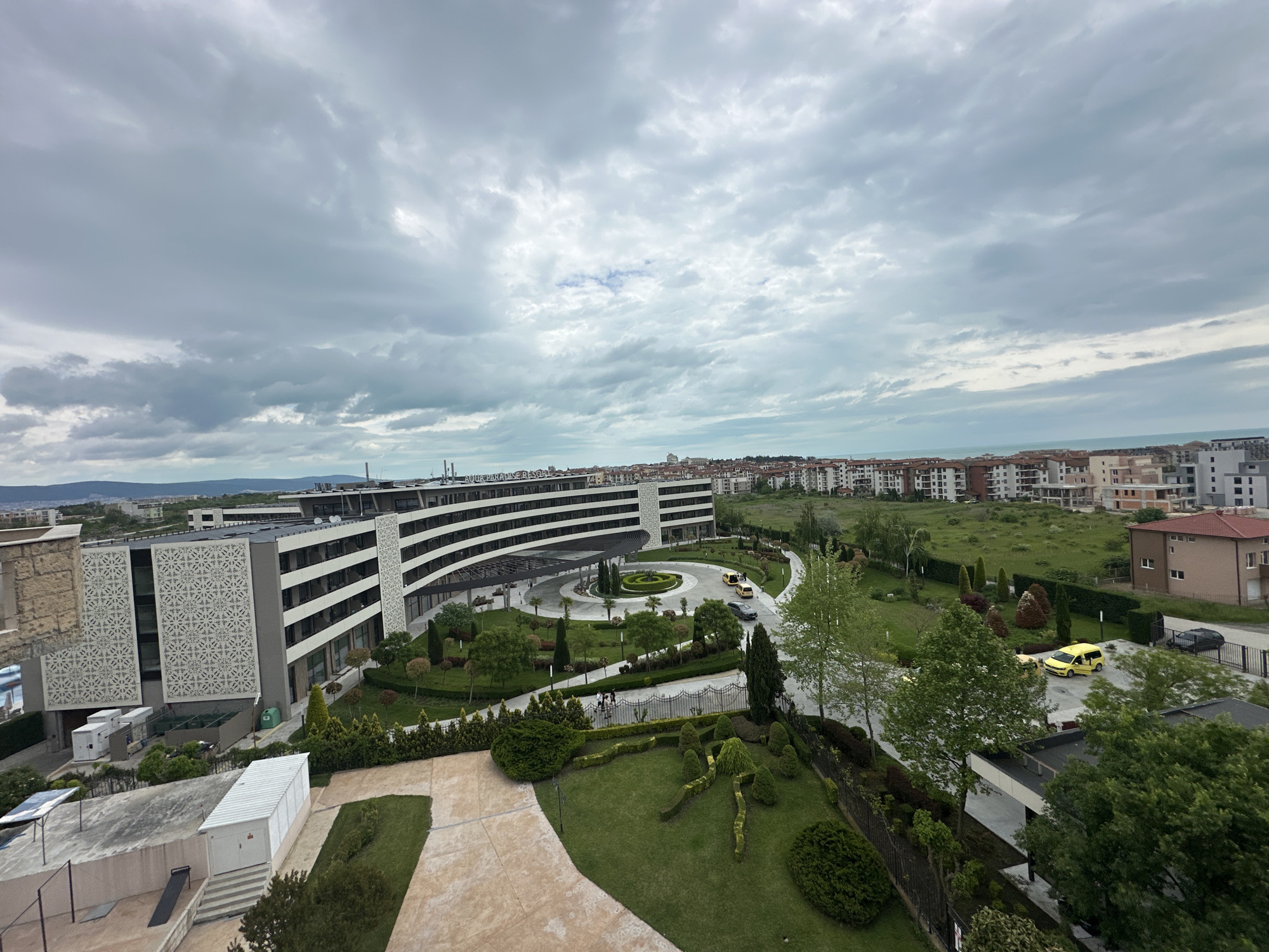 The image depicts a high-angle view in Ravda, Bulgaria. The focal point is a modern, curved hotel complex with a landscaped roundabout entrance. A garden featuring topiaries lies in the foreground. Several people are visible walking along a path near the hotel entrance. Beyond the hotel, the scene extends to residential buildings with terracotta roofs. The sky is overcast with gray clouds. A yellow vehicle is parked near a road in the middle ground.