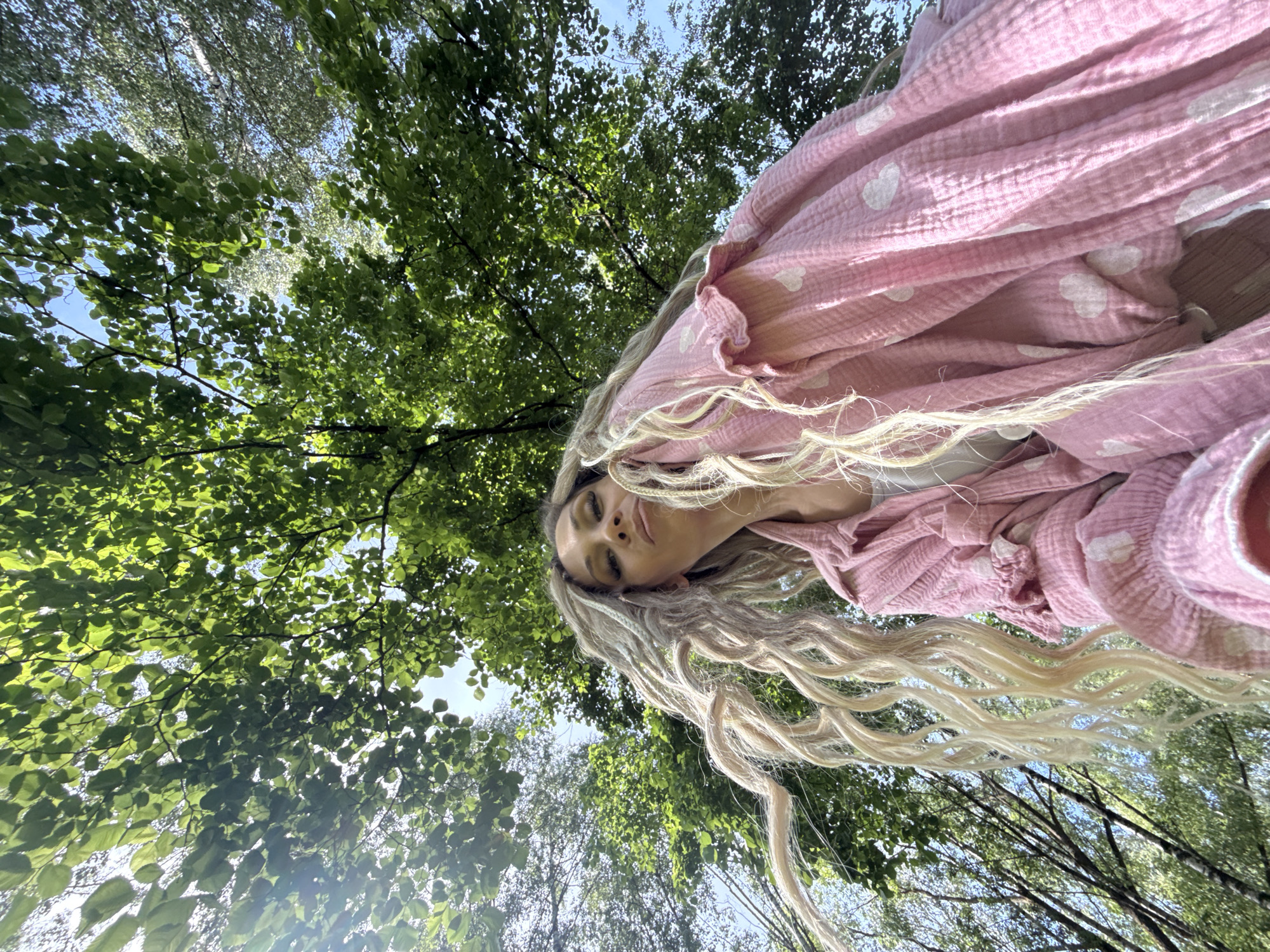 The image was captured in Rusanovo, Russia. It is a low-angle outdoor shot looking upwards into a dense canopy of green deciduous trees against a clear blue sky, with bright sunlight filtering through the leaves. An adult female is positioned in the lower half of the frame, looking down towards the camera with a slight pout. She has long, voluminous blonde hair, styled with numerous textured strands or dreadlocks, flowing downwards and to the left. She is wearing a loose-fitting, light pink long-sleeved shirt or jacket made of a textured fabric, patterned with small white hearts. A light-colored undershirt is partially visible beneath.