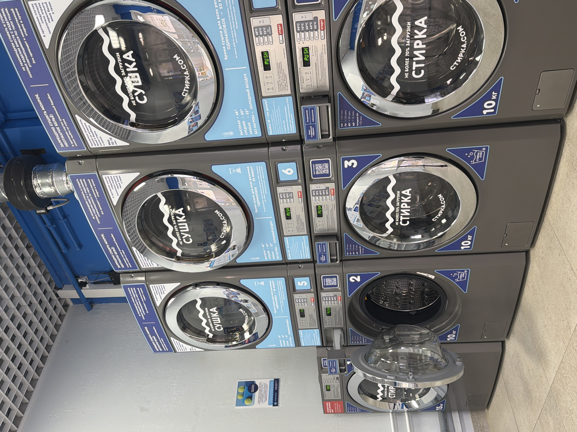 This is a photograph of a self-service laundry facility located in Samara, Russia. The scene captures multiple rows of stacked washing machines and dryers. The machines are predominantly grey with prominent blue accents and labeling. Many of the machines display Russian text, indicating operational instructions and brand information.
In the foreground, a washing machine with its door ajar reveals a stainless-steel drum. The adjacent machines are closed and appear to be operational or ready for use. The dryers are stacked above the washing machines. The overall environment suggests a clean and organized commercial space, with a tiled floor and a grid-like ceiling with visible lighting fixtures and ventilation systems.
On the wall to the left, a blue poster with images of yellow tennis balls and informational graphics is visible. The lighting is bright and even, typical of an indoor commercial setting. The perspective of the photograph is from a slightly low angle, looking up at the stacked machines. There are no people visible in the image.