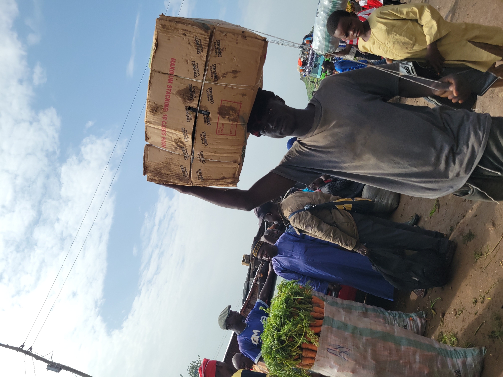 The image depicts an outdoor market scene with several individuals. In the foreground, a young Black male is carrying a large cardboard box stacked with other boxes on his head. The cardboard box has red lettering that reads "MAXIMUM STACKING: 10 CASES HIGH" and "NASCO CORNFLAKES". He is wearing a grey t-shirt and dark pants. To the left of him, a Black man wearing a blue t-shirt with "GM" on it and a green cap is interacting with another Black man in a blue traditional robe. Between them is a large sack filled with carrots. Further in the background, other people are visible, some carrying goods. The ground is dirt, and there are scattered green leaves and debris. The sky is partly cloudy with some blue patches. A power pole with wires is present in the upper left portion of the image. The overall setting suggests a bustling market environment in an urban area, likely in Africa, given the attire of the individuals and the types of goods displayed.