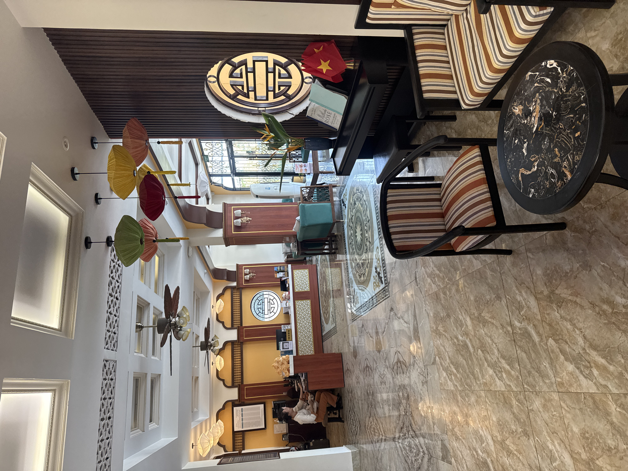 The image depicts the interior of a hotel or lobby in Đà Nẵng, Vietnam. The scene is viewed from an elevated perspective. The floor is covered in tan marble tiles. Near the front is a front desk with 2 women working at the desk. The interior design includes white walls, ceiling fans, and hanging decorative umbrellas. There is a seating area with striped armchairs and a round table with a black and gold marble top. A grand piano sits near the seating area with the Vietnamese flag and a book atop. A decorative sign with the same pattern as the umbrellas.