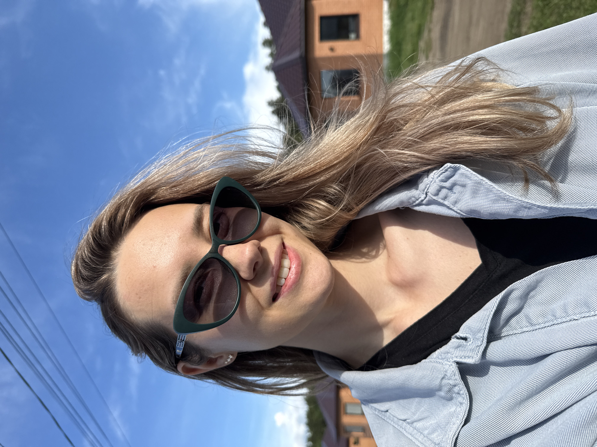 A young woman is captured in a rotated, close-up portrait, smiling broadly at the viewer in an outdoor setting. She has long, wavy, light brown hair, brightly illuminated by direct sunlight. She is wearing dark green, cat-eye shaped sunglasses and small, star-shaped earrings. Her mouth is open in a wide, joyful smile, revealing her white teeth. She wears a light blue, possibly chambray or denim, button-up shirt over a black top.
The background shows a clear, bright blue sky with a few wispy white clouds, indicating sunny weather. A reddish-brown building with dark windows, likely a house, is visible in the upper background, along with green vegetation. Power lines are visible against the sky on the left side of the frame, suggesting a residential or semi-urban area. The strong sunlight and clear sky indicate it is daytime, possibly midday. The woman appears happy and relaxed. The location is Chaikovsky, Russia. No visible text is present in the image.