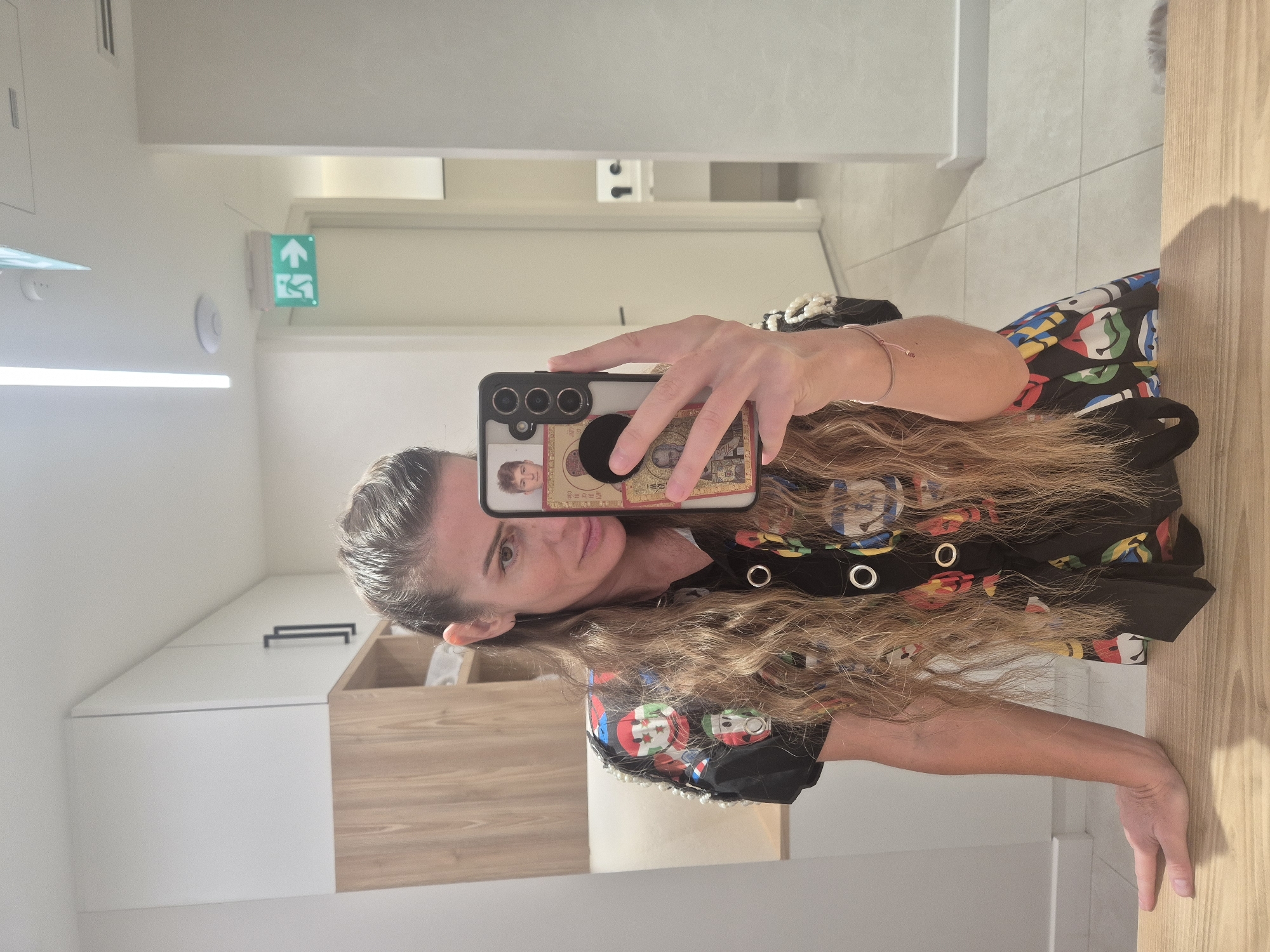 An image, rotated 90 degrees clockwise, depicts a woman taking a mirror selfie indoors. The woman, seen from the chest up, has long, wavy, light brown hair flowing over her shoulders. She looks towards the camera with a neutral to slightly smiling expression. Her left hand holds a black smartphone horizontally, featuring a triple camera array and a PopSockets grip. The phone case has a small photo of a young man and what appears to be an ornate, possibly religious, artwork or icon. On her left wrist, she wears a slender, light pink bracelet.
She is wearing a black top or dress adorned with a pattern of colorful, cartoon-like faces or lips in red, blue, green, and yellow, along with circular silver eyelets on the front. Pearls or white beads are visible on the sleeve of her right arm, which rests on a light wooden surface in the foreground.
The background shows a clean, modern interior with light-colored walls. To the left, a bright horizontal light fixture is visible. Further back, an emergency exit sign with an illuminated green background displays an arrow pointing upwards and a running person symbol. The interior features white and wooden cabinetry or shelving units. The lighting appears to be artificial, typical of an indoor setting.