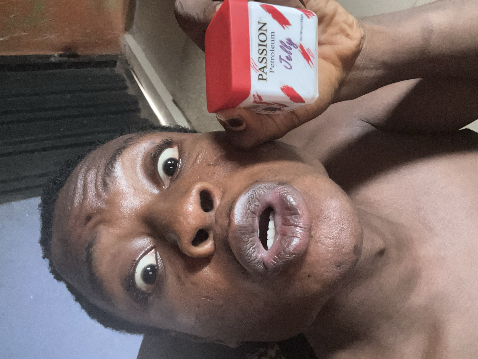 A person with brown skin is holding a container of Passion Petroleum Jelly in their right hand. The person's mouth is open, and their eyes are wide. The background is out of focus. The location of the city is unknown.