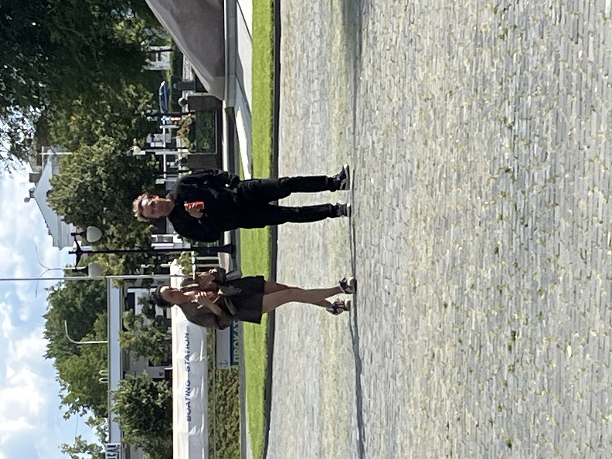 Two people, a woman and a man, are standing in a city square paved with cobblestones. The woman, on the left, is wearing a dark brown jacket, a short dark skirt, and sneakers. She is holding a drink and appears to be talking or interacting with the man. The man, on the right, is wearing a black hoodie and dark pants. He is holding a red can, likely a beverage, and is looking towards the camera. Behind them, there is a grassy area with trees and buildings. A white banner with the words "BOATING STATION" is visible in the background. The overall scene suggests a casual outdoor setting in a city.