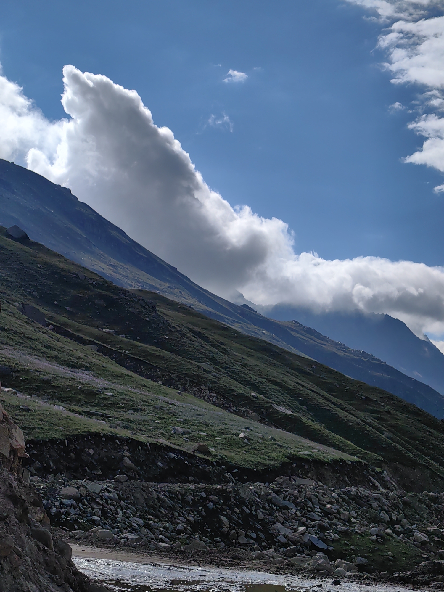 A dramatic, low-angle landscape shot captures the rugged beauty of a mountain pass in Bashisht, India, under a dynamic sky. The scene is dominated by steep, grassy slopes that rise diagonally from the lower left to the upper right of the frame. These slopes are dotted with small, scattered rocks and patches of low-lying purple wildflowers, creating a textured, verdant carpet. A sharp, dark embankment cuts across the mid-ground, revealing a layer of soil and a cascade of larger stones and rocks. Below this embankment, a rough, muddy track, possibly a road, winds through the foreground, with puddles of water reflecting the sky.
The sky above is a vibrant blue, punctuated by dramatic, white cumulus clouds. One particularly striking cloud formation in the upper left resembles a sharp, pointed shape, possibly a bird or a spearhead. Another line of fluffy clouds stretches across the upper right, partially obscuring distant, hazy mountain peaks. The overall impression is one of wild, untamed nature, with the formidable presence of the mountains and the ever-changing sky setting a majestic tone. There are no people or animals visible in the frame. The lighting suggests a bright, possibly midday, sun, casting subtle shadows on the terrain.