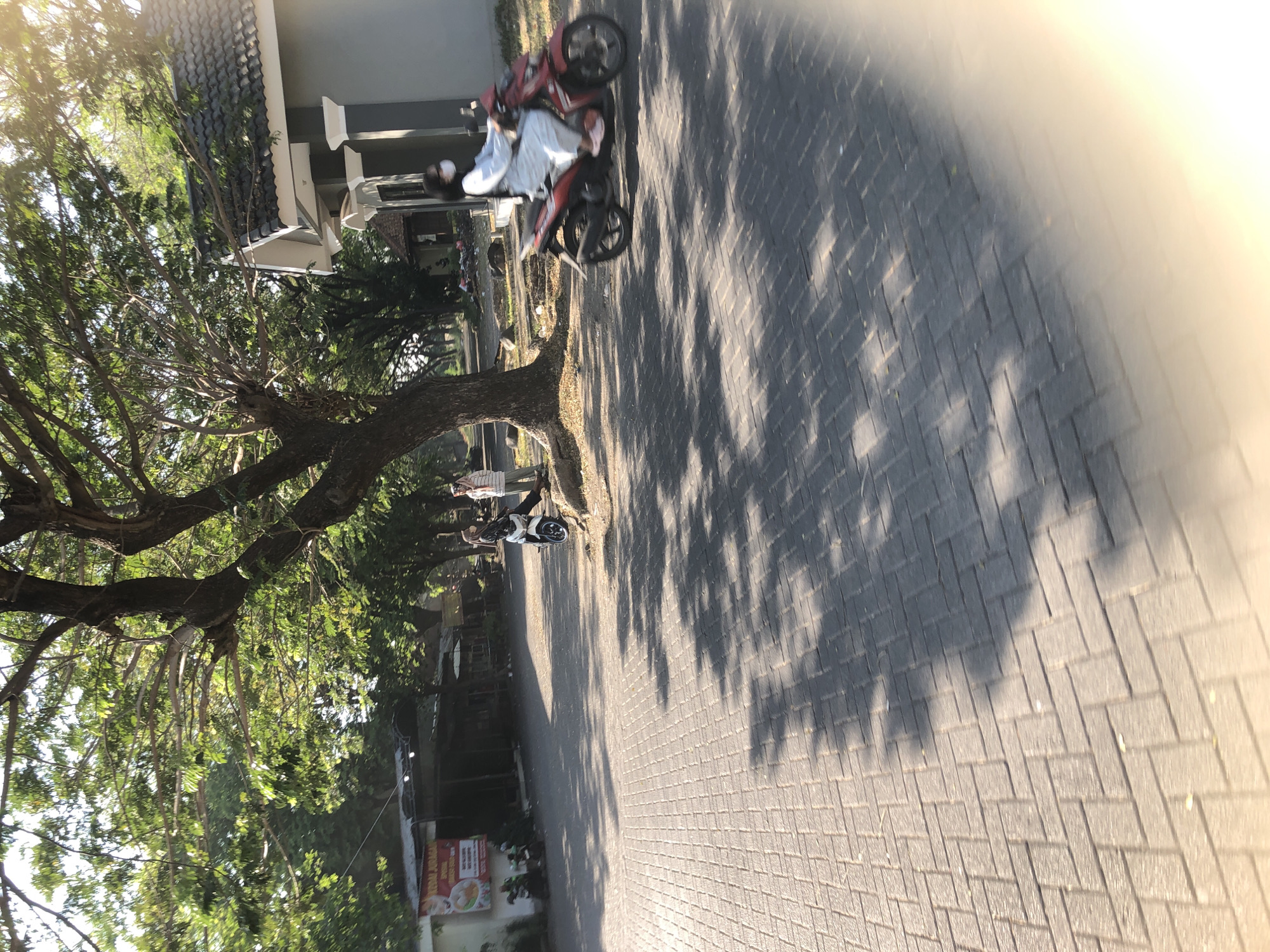 The image shows a paved area in front of a building in Boro, Indonesia. On the right side, a person is riding a red motorcycle, wearing a white outfit and a face mask. In the background, there are trees and another building. To the left, there is a banner that appears to be advertising food. Two other individuals are standing near a white motorcycle on the left side of the image. The ground is paved with interlocking bricks, and shadows from trees are cast across the surface. The scene is illuminated by sunlight, suggesting it is daytime.