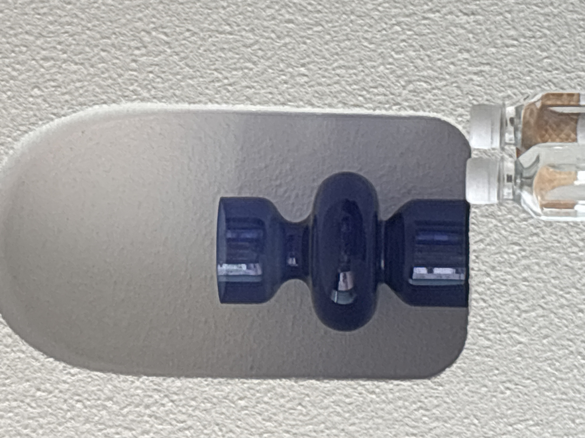 A close-up shot shows a dark blue, dumbbell-shaped coat hook mounted on a textured, light gray or white wall. The hook is affixed to the wall by a clear adhesive strip that extends beyond the hook's base. At the right edge of the frame, part of a curtain rod is visible, with clear plastic components attaching it to the hook. The scene is well-lit. I cannot determine the specific location in Puebla, Mexico, from the given image.