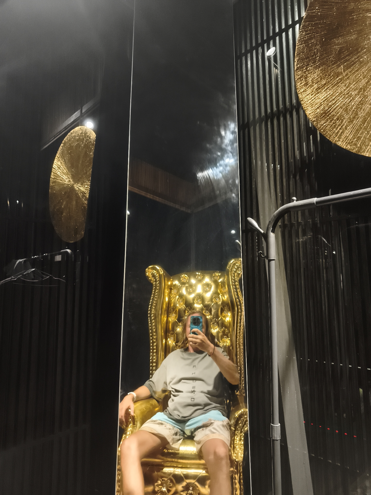 A person is seated in a gilded throne in what appears to be a dimly lit indoor space. The throne has a high, tufted back and ornate arms. The person is wearing a gray t-shirt and patterned shorts, and is holding a blue smartphone up to take a selfie in a tall, full-length mirror that reflects the scene. Behind the throne, a dark wall with vertical fluting is visible, adorned with two large, circular gold decorative elements. To the right of the throne and mirror, a metal clothing rack with a grey pole is present. The overall aesthetic suggests a luxurious or dramatic setting. Based on the limited visual information available in the image, it is not possible to determine the specific location within Baan Mae Nam, Thailand.