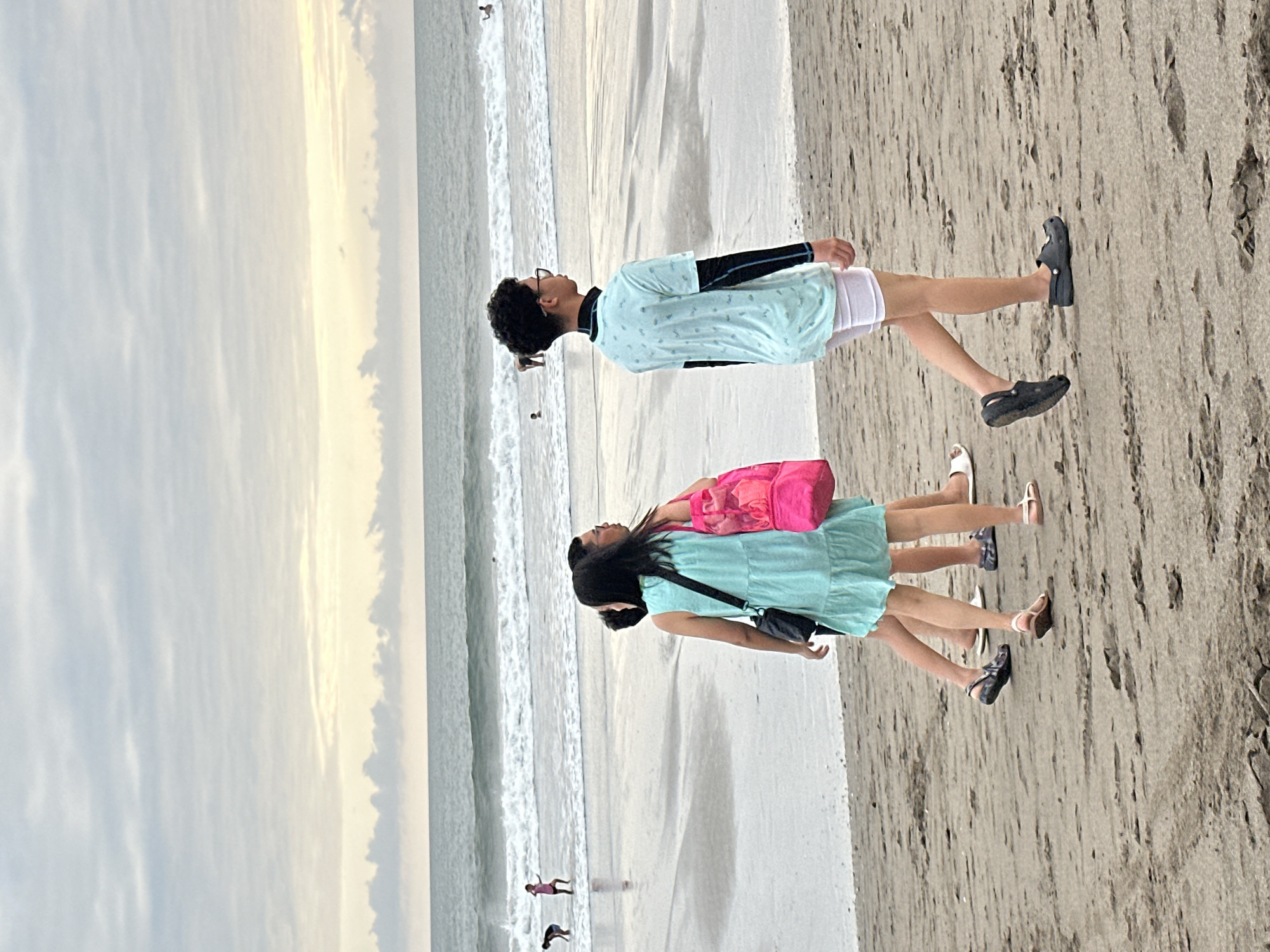A group of four people are walking on a sandy beach in North Kuta, Indonesia. The sky is overcast with a pale yellow glow near the horizon. The ocean has moderate waves. In the foreground, two children are walking ahead, followed by a teenage girl carrying a pink bag and a teenage boy wearing glasses. In the background, several other people are visible in the water, some appearing to be swimming or surfing. The sand is damp and shows some debris.