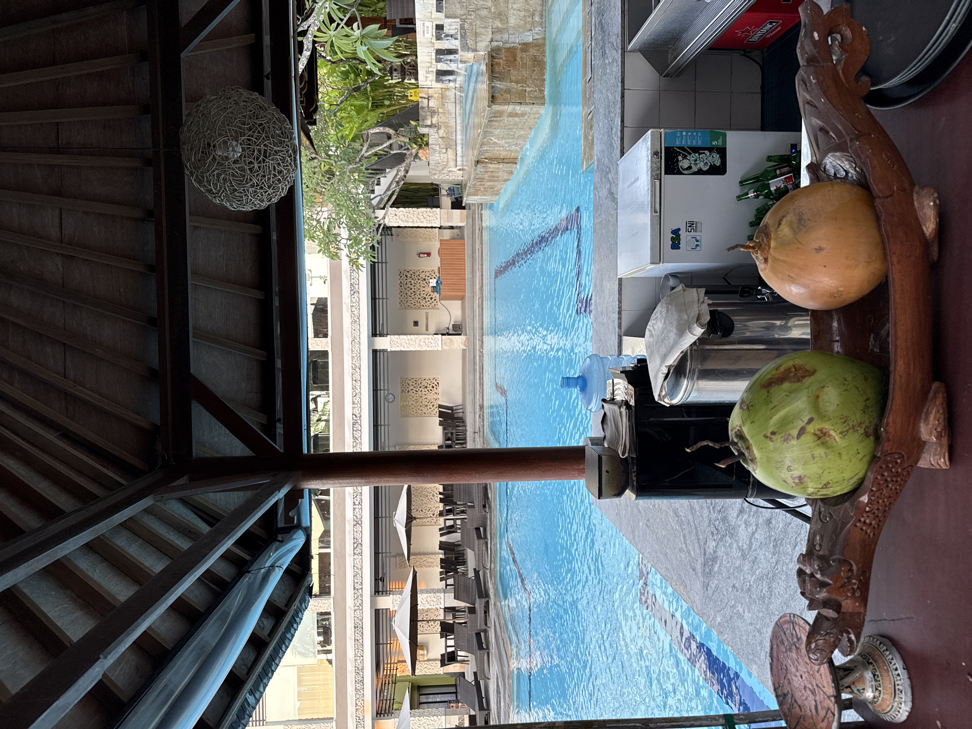 This image captures a vibrant poolside scene, likely at a resort or hotel in Kuta Selatan, Indonesia, viewed from under a shaded, open-air structure. The perspective is taken from a bar or lounge area looking out onto a swimming pool.
In the foreground on the right, two coconuts, one green and one yellowish-orange, rest on an ornately carved wooden support that appears to be part of a counter. Behind them, a white refrigerator is visible, partially filled with green bottles, likely beverages. Above the refrigerator, a red crate or box displays the text "BINTANG." The refrigerator itself has "RSA" and "SNI" labels. A large blue water dispenser bottle is also on the counter.
The central feature of the scene is a spacious swimming pool with clear blue water and dark lane markers on its bottom. Beyond the pool, rows of lounge chairs and tables, some with white umbrellas, are arranged on a paved deck, indicating a sunbathing and relaxation area. In the background, multi-story resort buildings with balconies and decorative architectural elements are visible, interspersed with lush green trees and tropical foliage.
Overhead, a distinctive spherical light fixture, appearing to be made of woven natural fibers, hangs from a wooden beam. The intricate wooden roof structure and beams of the shaded area are prominent in the upper left and lower left portions of the image. The scene is illuminated by bright daylight, suggesting a clear and sunny day. There are no people visible, but the setup implies a leisure or vacation setting.