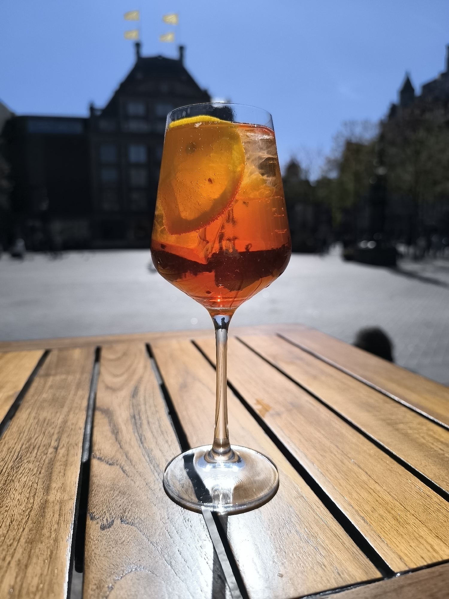 A medium shot features an Aperol Spritz in a wine glass, positioned on a wooden table with slats. The glass is filled with the orange-hued cocktail, garnished with orange slices. The setting appears to be an outdoor café or patio.
In the background, blurred, is a city square in Amsterdam, Netherlands. Buildings with intricate architectural details are visible, including the back of the Palace on Dam Square with its distinctive yellow flags. The sky is a bright blue, suggesting a sunny day. A few blurred figures can be seen in the square, indicating public activity.