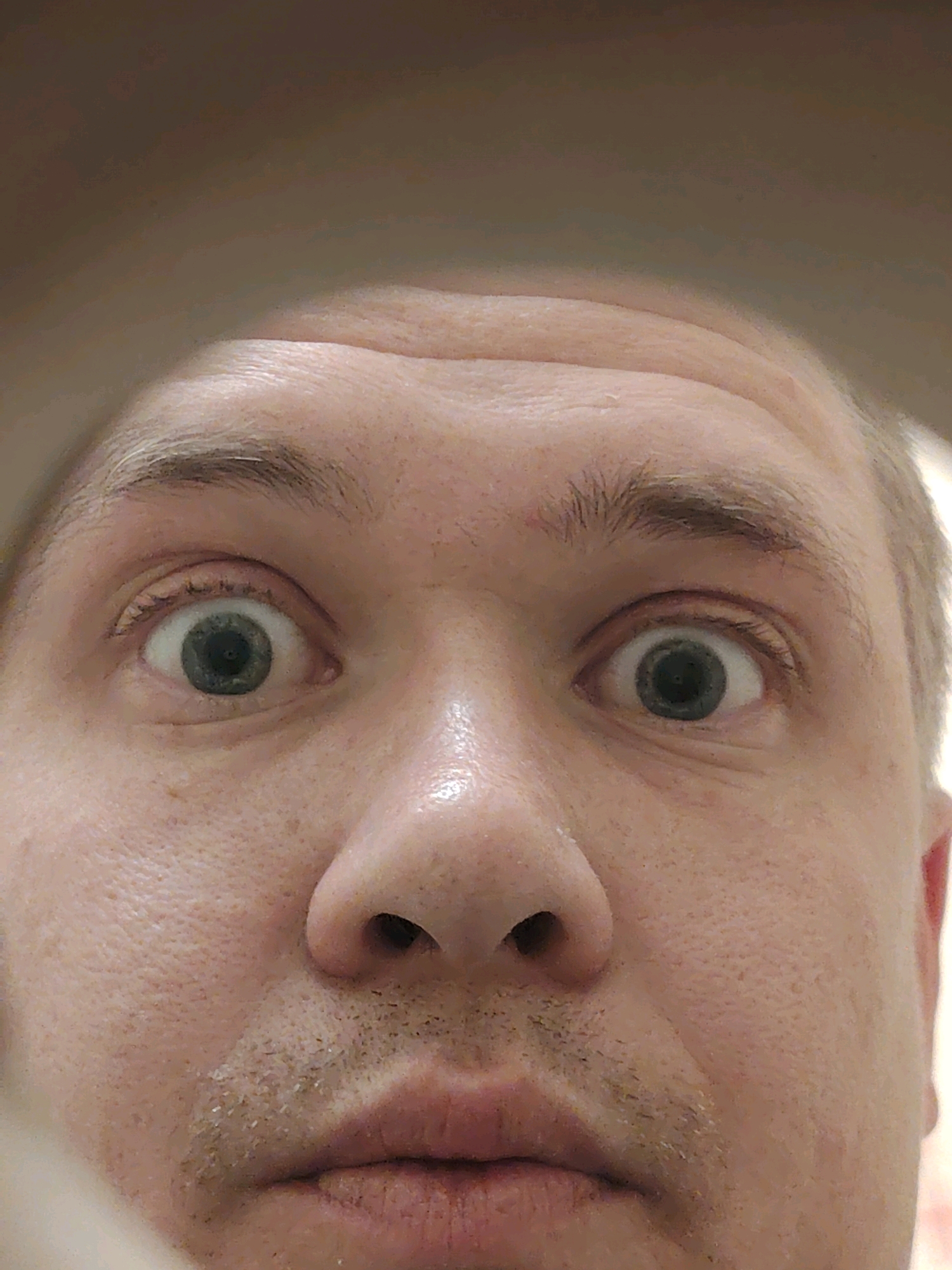 This is a close-up, selfie-style photograph of a person's face, focusing on their eyes, nose, and mouth. The individual has light-colored skin with visible pores and a faint stubble on their upper lip and chin. Their eyes are wide open and a light gray-green color, with visible eyelashes and eyebrows. The lighting suggests an indoor environment, possibly a bathroom or bedroom, given the soft, diffused light. There are no other people or objects clearly visible in the frame. The background is out of focus and appears to be a neutral, muted color, possibly a wall or ceiling. This image does not contain any identifiable landmarks or elements that would suggest a specific location within the city of Chaikovsky, Russia.