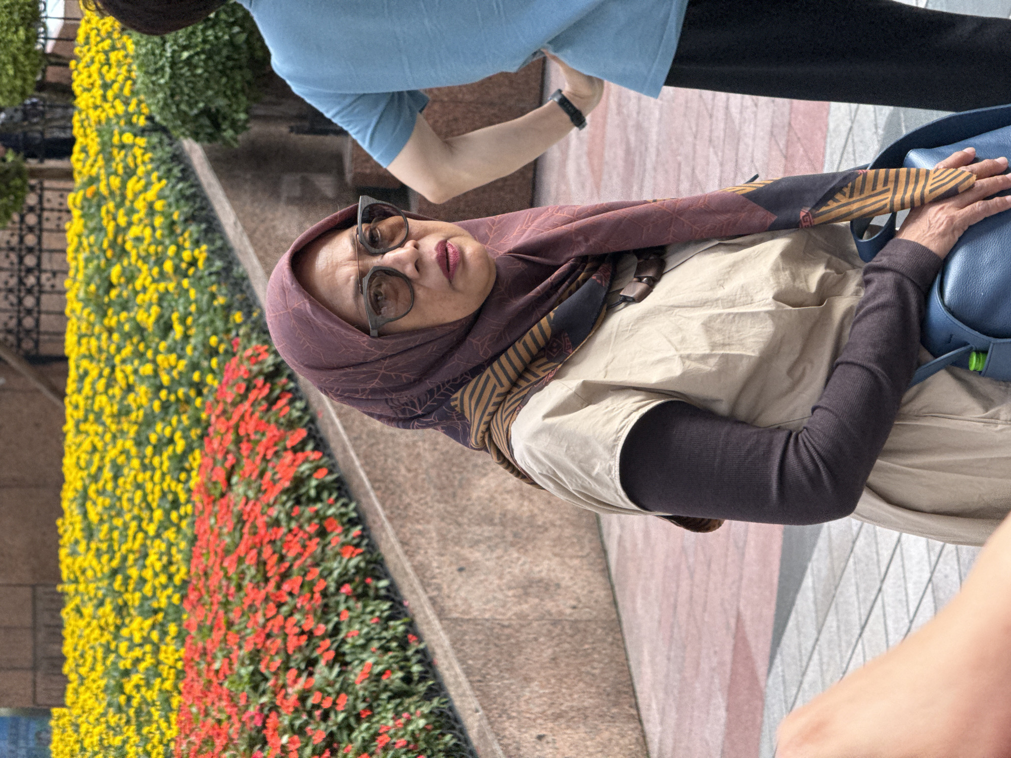 An eye-level shot captures a woman in a vibrant outdoor setting. She is positioned towards the center-right of the frame, looking directly towards the viewer with a neutral to slightly serious expression. She wears a patterned maroon and brown hijab, dark-rimmed glasses, a light tan top, and a dark, long-sleeved undershirt. Her hands rest gently on a blue handbag.
Behind her, a striking landscape feature consists of two parallel rows of densely planted flowers: one of bright yellow blooms and another of vivid red flowers, adding a burst of color to the scene. These flower beds run alongside a paved pathway.
In the upper background, part of another person is visible, wearing a light blue t-shirt and dark clothing, with a watch on their wrist. The arm and partial body of a third person are visible in the lower right foreground. The lighting suggests it is daytime. The overall impression is a public park or pedestrian area in Yuzhong District, China, characterized by well-maintained flora and urban pathways. No discernible text is visible in the image.