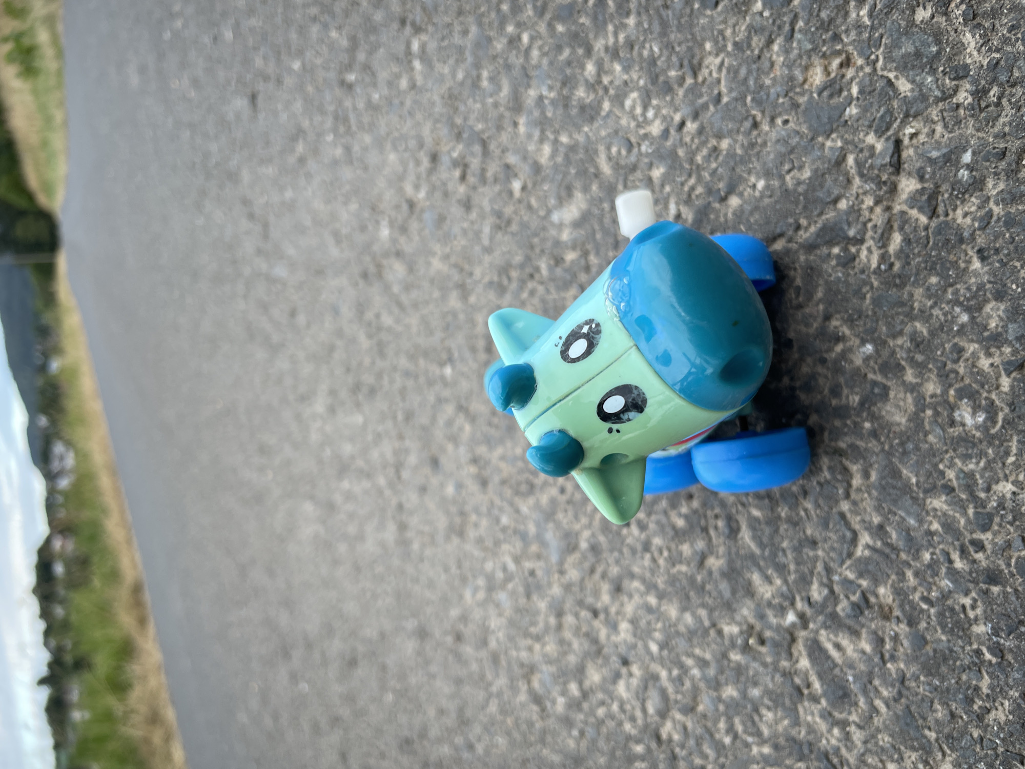A small, toy airplane lies on its side on a textured asphalt surface. The toy is primarily light green with a teal blue cockpit and tail. It has large, cartoonish black and white eyes with visible pupils. The airplane has two small, teal blue propellers or wings protruding from the top of its cockpit and two teal blue wheels attached to its underside. A white piece of plastic extends upwards from the cockpit. The asphalt is dark gray with visible aggregate. To the left of the asphalt, a grassy verge slopes down towards what appears to be a body of water or a ditch, with green grass and some brown vegetation. A faint reflection can be seen in the water. The scene is outdoors, likely in a rural or roadside setting, given the grassy verge. The lighting suggests it is daytime. There is no visible text in the image. The image is taken from a slightly elevated angle, looking down at the toy.