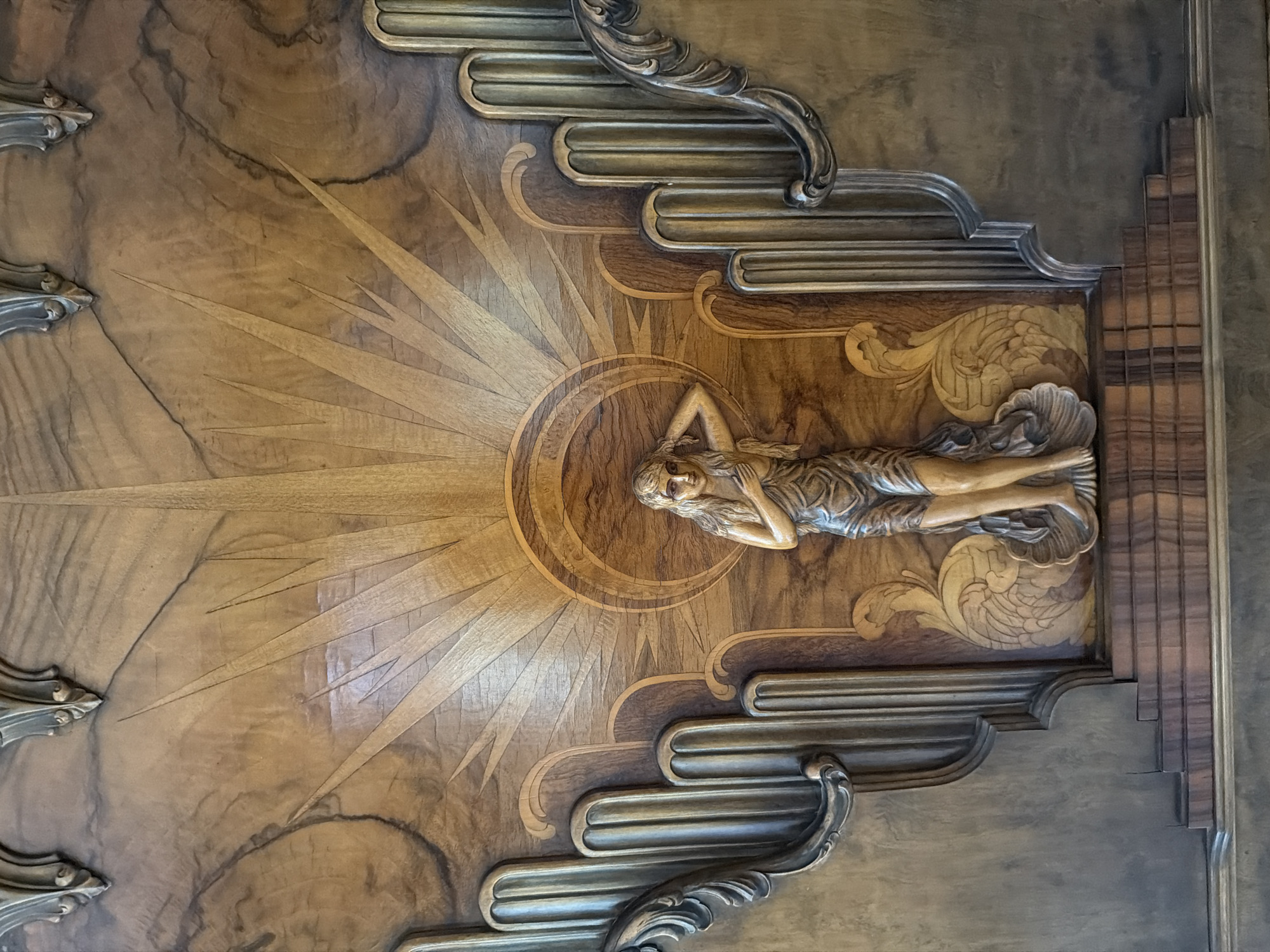 The image presents a horizontal close-up of an ornate wooden panel. The panel appears to be made of different types of wood, and its central design depicts a woman reclining horizontally atop a sunburst pattern. The sunburst emanates from a circular area behind the woman. The woman is carved in relief and appears to be nude or partially draped. Her hands are positioned near her face. The design incorporates elements resembling stylized waves or clouds above and below the sunburst. Metallic-toned, stylized carved details adorn the edges. The background wood has a darker grain, and the right edge of the panel displays vertical banding. This panel is located within the Paramount Theatre in Oakland, California.