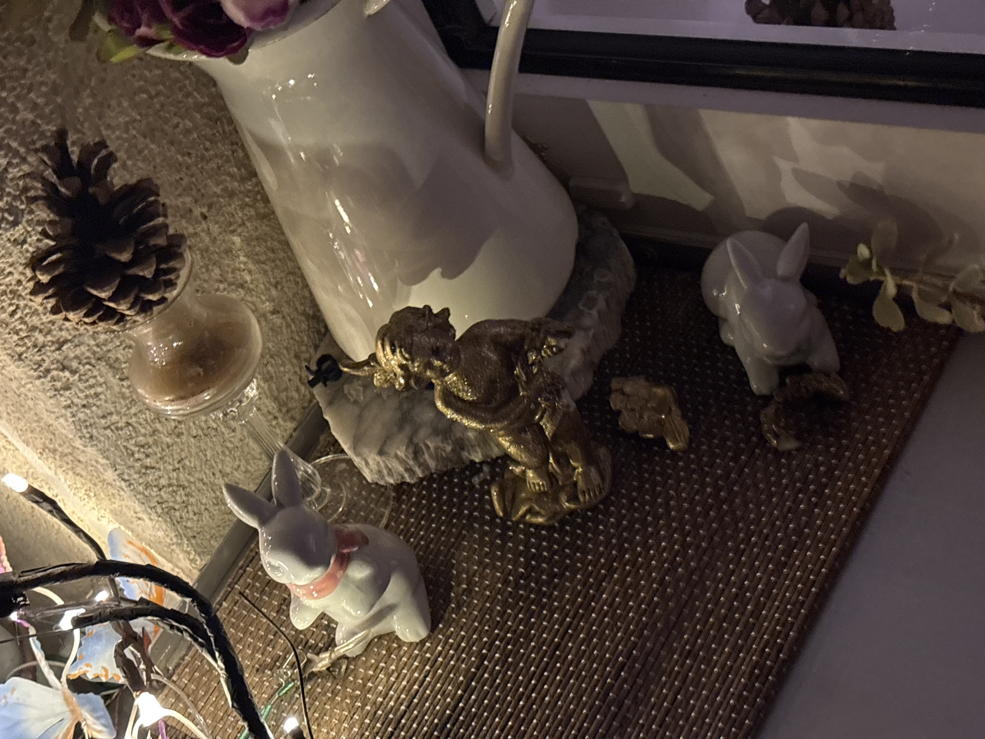 The image shows a still life arrangement with various decorative objects. In the foreground and midground, there are several ceramic rabbit figurines, one of which is white with a pink collar. A gold-colored figurine, possibly a cherub or angel, is positioned on a piece of marble. A pine cone rests on a textured surface next to a clear glass vessel containing a light brown substance. In the background, a white ceramic pitcher holds purple and pink flowers, and a string of small lights is visible in the lower left corner. The objects are placed on a woven mat. There are no people visible in the image. The scene does not provide any contextual information to determine its location in Osterode am Harz, Germany, or any other specific city.