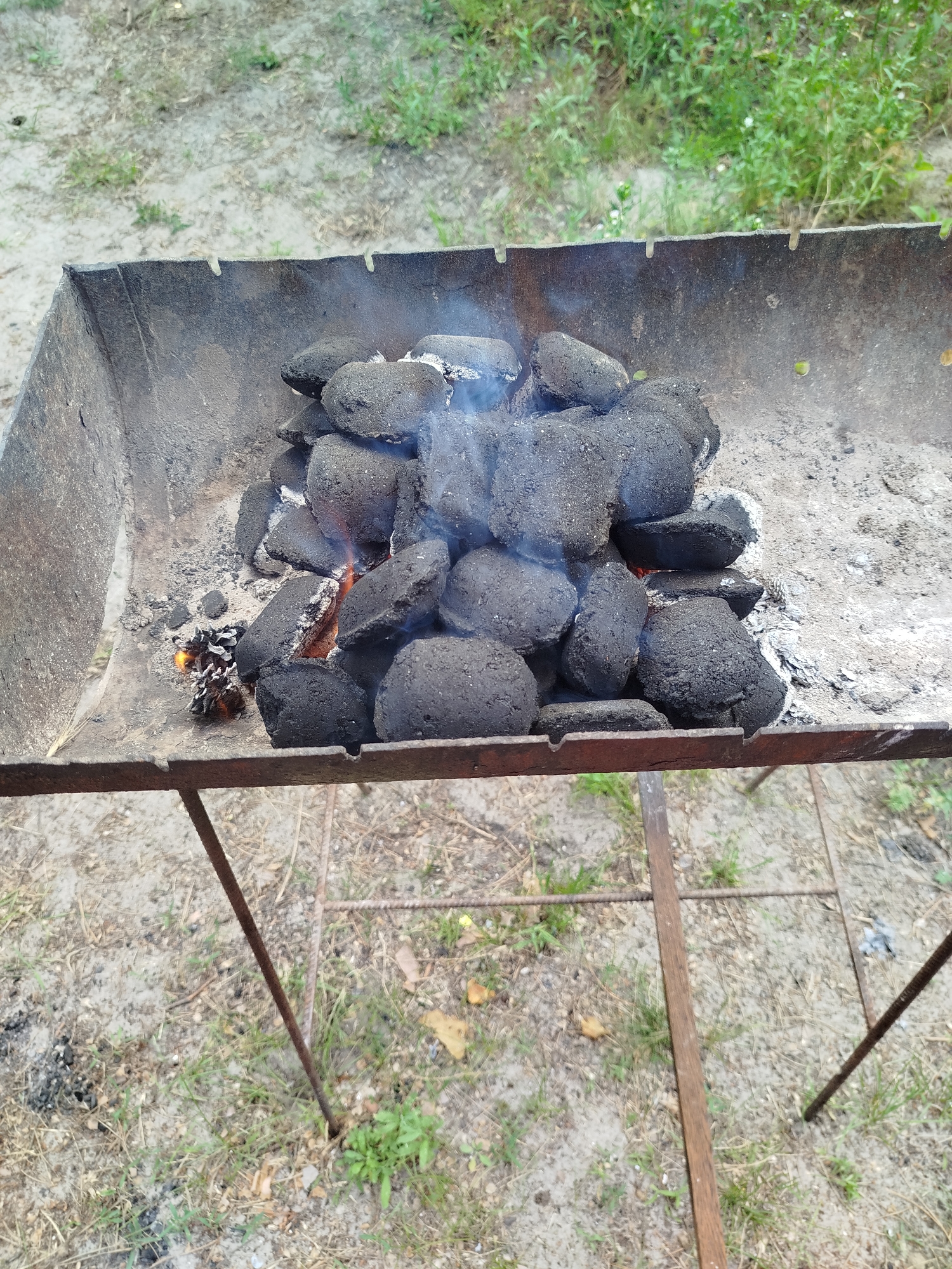An overhead shot shows a charcoal grill filled with hot coals. The grill is a simple metal box with legs, and it sits on the ground in an outdoor area with sandy soil and some sparse vegetation. The coals are piled high in the center of the grill, and some are glowing red, indicating they are hot. Smoke is rising from the coals, and there are a few flames visible. The grill appears to be in use, possibly for a barbecue.