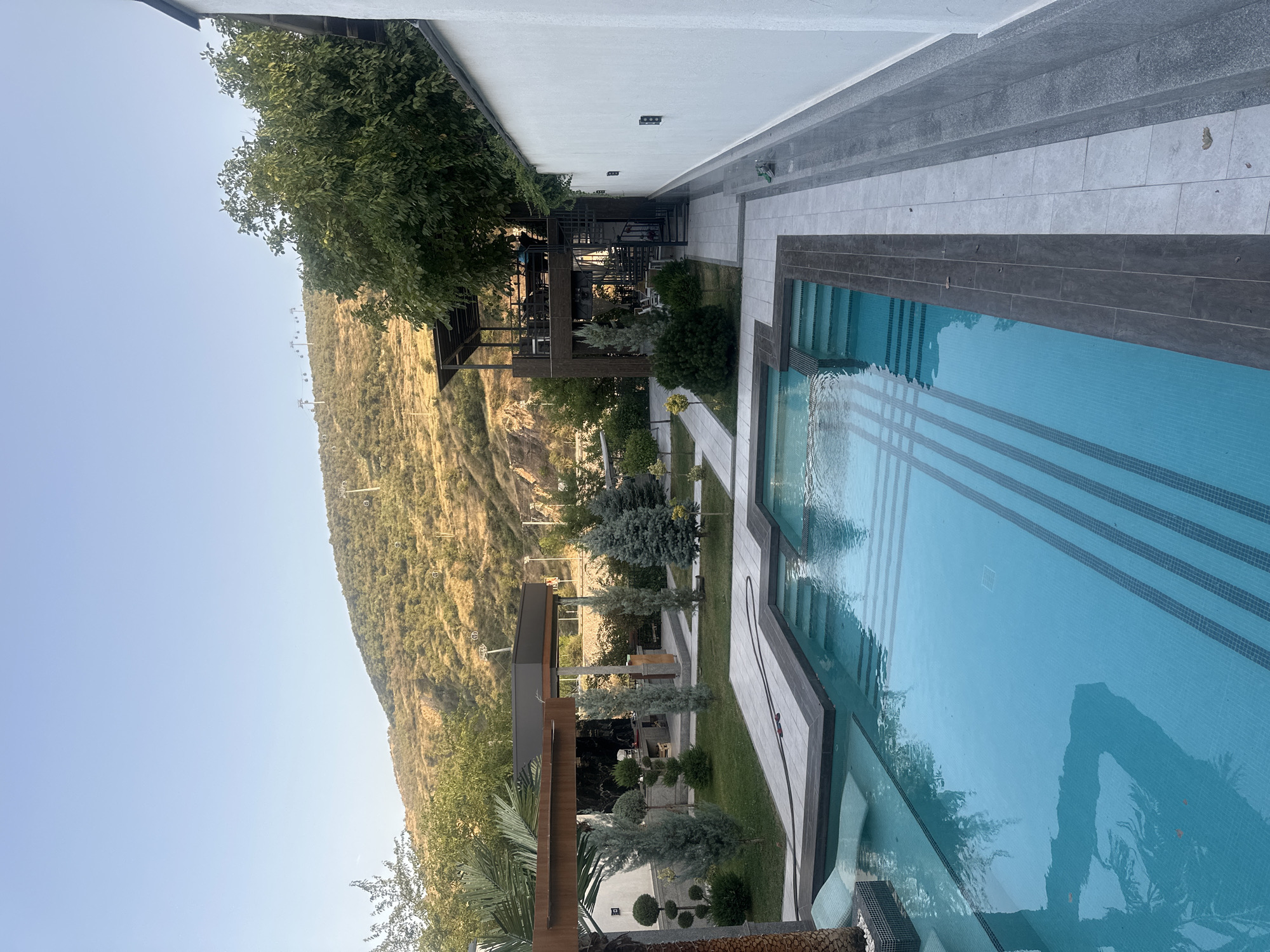 An exterior wide shot captures a luxury outdoor pool area with a mountainous backdrop in Charvak, Uzbekistan. The foreground is dominated by a large, turquoise swimming pool featuring tiered steps and a checkered mosaic bottom. To the left of the pool, a white chaise lounge is partially submerged in a shallow section.
Beyond the pool, a meticulously landscaped garden with manicured trees and shrubs unfolds. A paved walkway with grey tiles leads through the garden. In the mid-ground, a multi-level wooden structure with a dark roof and an external spiral staircase is visible, with tables and chairs set up on the upper level.
The background consists of a dry, brown hillside covered with sparse vegetation, dotted with utility poles and what appear to be ski lifts extending to the summit. The sky above is a clear, pale blue. A white, textured wall with dark rectangular fixtures runs along the right edge of the image, framing the pool and garden area. The scene appears to be set during daylight, likely late afternoon, given the soft, warm light.