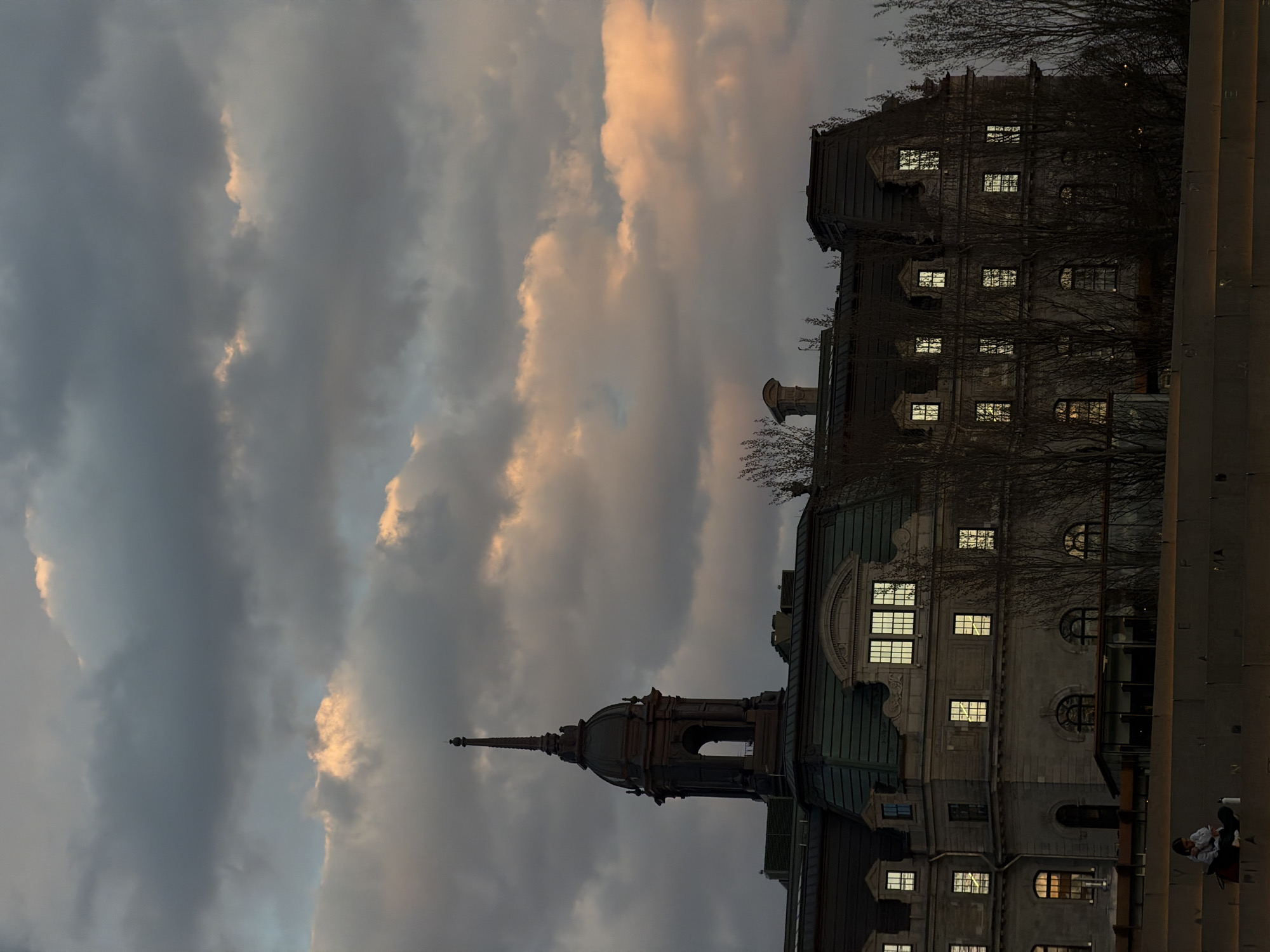 The image depicts a tall building in Montreal, Canada, seen from a low angle against a backdrop of a cloudy sky at sunset. The building, likely situated in Old Montreal, features multiple floors with lighted windows, suggesting it is dusk or twilight. Its architecture includes a prominent tower and intricate detailing. Bare tree branches partially obscure the building's facade. In the lower right corner, two individuals are visible, possibly tourists or locals observing the scene. The overall environment suggests a historic urban setting with a mix of architectural grandeur and a serene sunset atmosphere.
