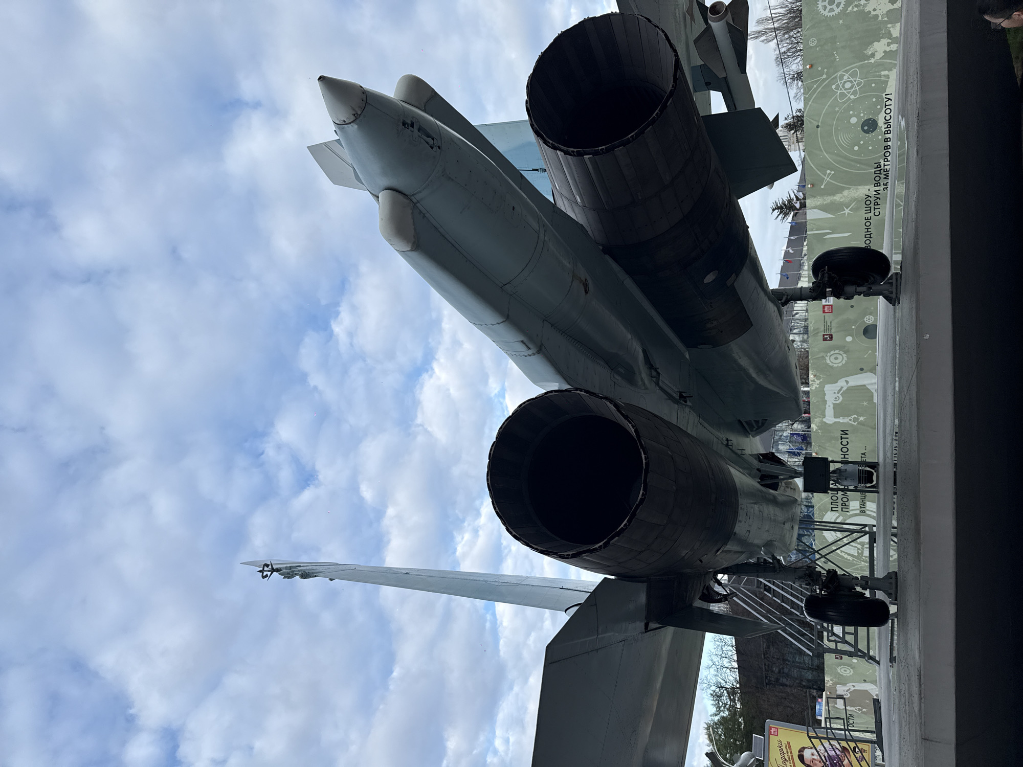 The image depicts the rear of a Su-27 fighter jet. The jet is light gray with two prominent engine exhausts. A tail fin is visible on the left. The background shows a cloudy sky.
The image was taken at the VDNKh park in Moscow, Russia. A panel behind the jet has some Russian text.