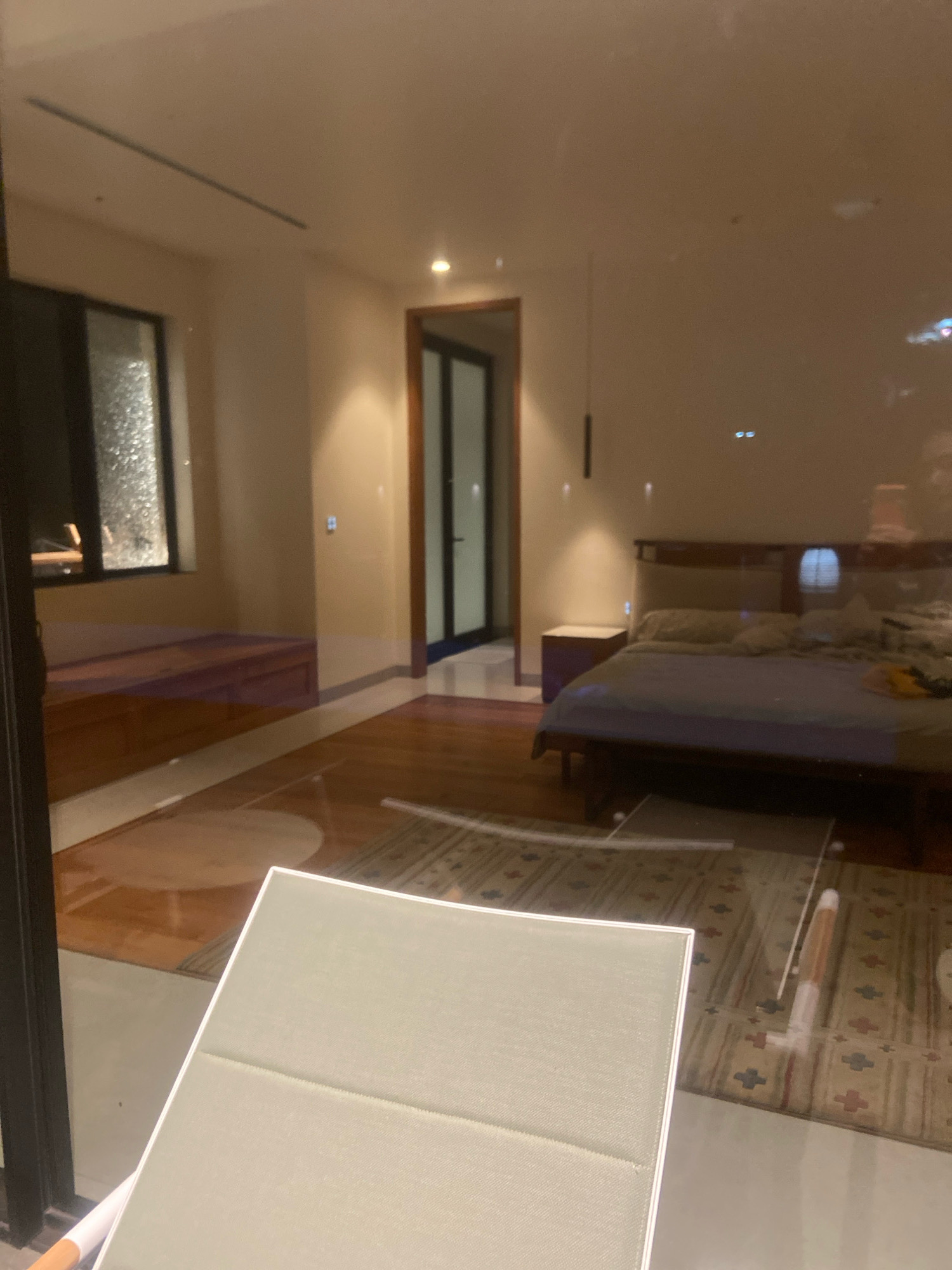 The image displays a reflection of a bedroom interior, likely captured from outside a glass-paneled door or window, in what appears to be a modern, well-lit residence. The bedroom features a large bed with a dark wooden frame and a upholstered headboard, covered with light-colored bedding and several pillows. A dark wooden nightstand with a single drawer is positioned beside the bed. The room has wooden floors and beige walls, with track lighting visible on the ceiling. A doorway leads to another part of the house, which is also visible in the reflection. A window with a frosted or textured glass is present on the left side of the bedroom, above a wooden bench or built-in seating area. A rug with a geometric pattern is spread across the floor in front of the bed. A light-colored chair or chaise lounge is partially visible in the foreground, suggesting the viewer's perspective. The overall impression is one of a clean, contemporary living space. The location in Ngaglik, Indonesia, is not directly evident from the visual content of the image, as there are no distinct Indonesian cultural or architectural markers.
