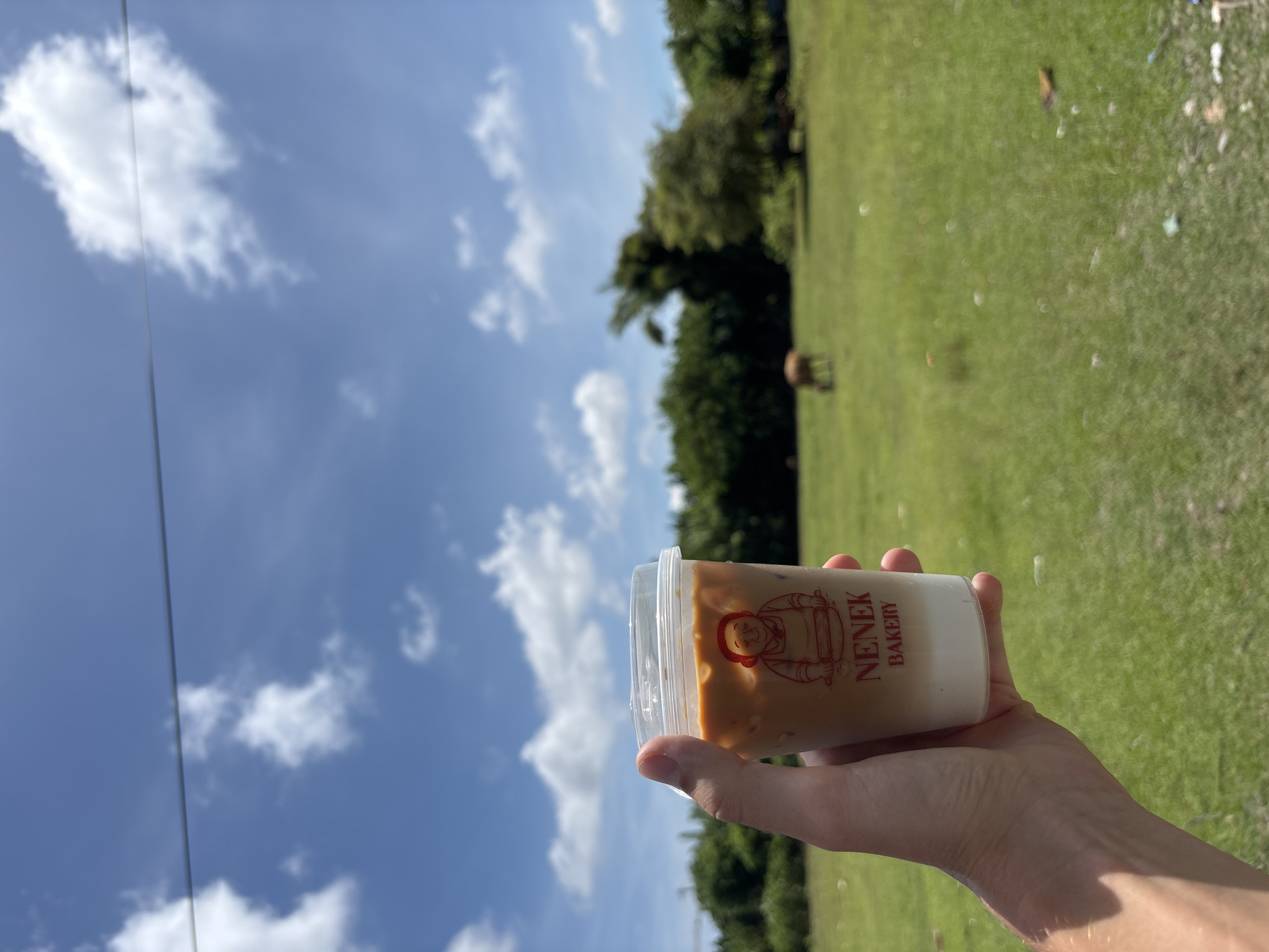 The image shows a hand holding a plastic cup containing a two-layered beverage. The cup has a red logo featuring a cartoon-like figure and the words "NENEK BAKERY". The beverage appears to be a mix of coffee or tea on top and milk on the bottom.
The background is a bright outdoor scene. Above is a blue sky with scattered white clouds. Behind the cup is a green field with trees in the mid-ground.
Given the branding on the cup, and as requested, the location of the image can be specified as Ungasan, Indonesia.