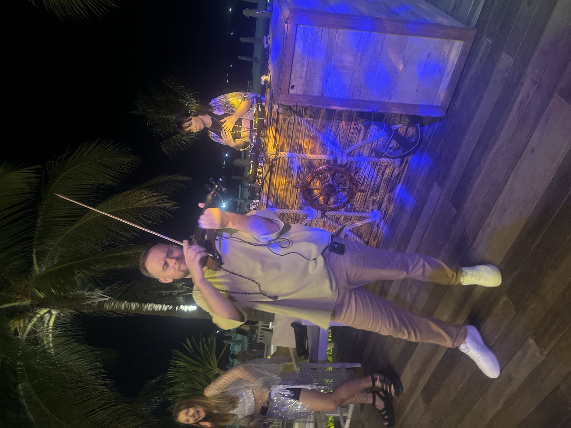 A man in a light-colored t-shirt and beige pants plays a violin on a wooden deck at night, illuminated by blue and orange lights. He wears a beaded necklace and has a small device clipped to his pants. To his left, a woman in a sparkly silver outfit laughs and poses. Behind the violinist, a DJ with headphones stands at a wooden booth decorated with nautical elements like a ship's wheel and rope, operating DJ equipment. The background features palm trees against a dark sky and glimpses of beach chairs and the sea. The scene appears to be an outdoor event, possibly in a resort area in Đà Nẵng, Vietnam, given the tropical foliage and beach setting.