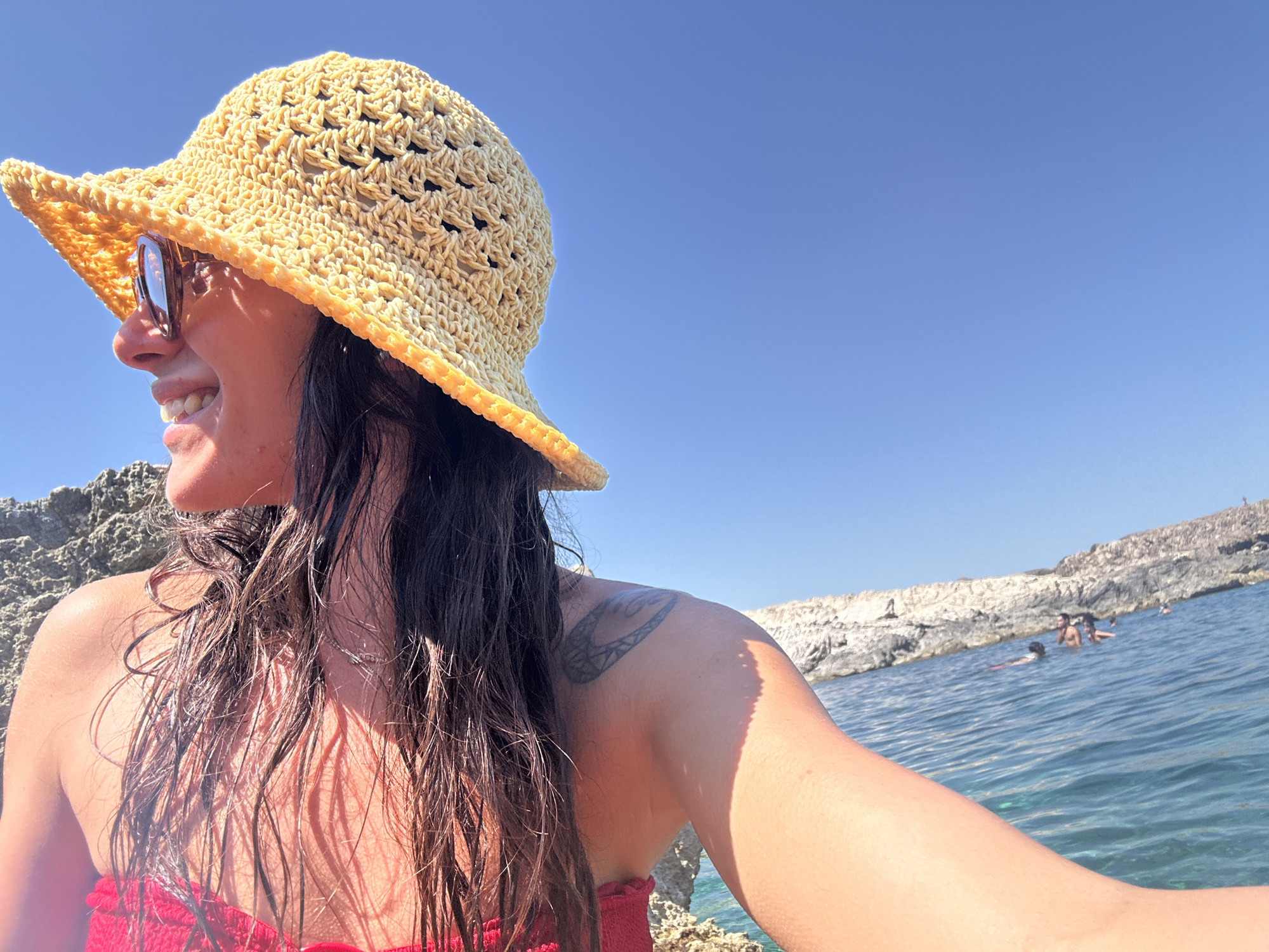 A woman is pictured close up from the chest up, wearing a yellow crocheted hat and sunglasses. She has dark, wet hair and a red bikini top. A tattoo is visible on her upper arm. She is smiling and looking to her left. The background shows the ocean with a rocky shoreline and other people swimming. The image appears to be taken at Cala Lliteres, Spain.