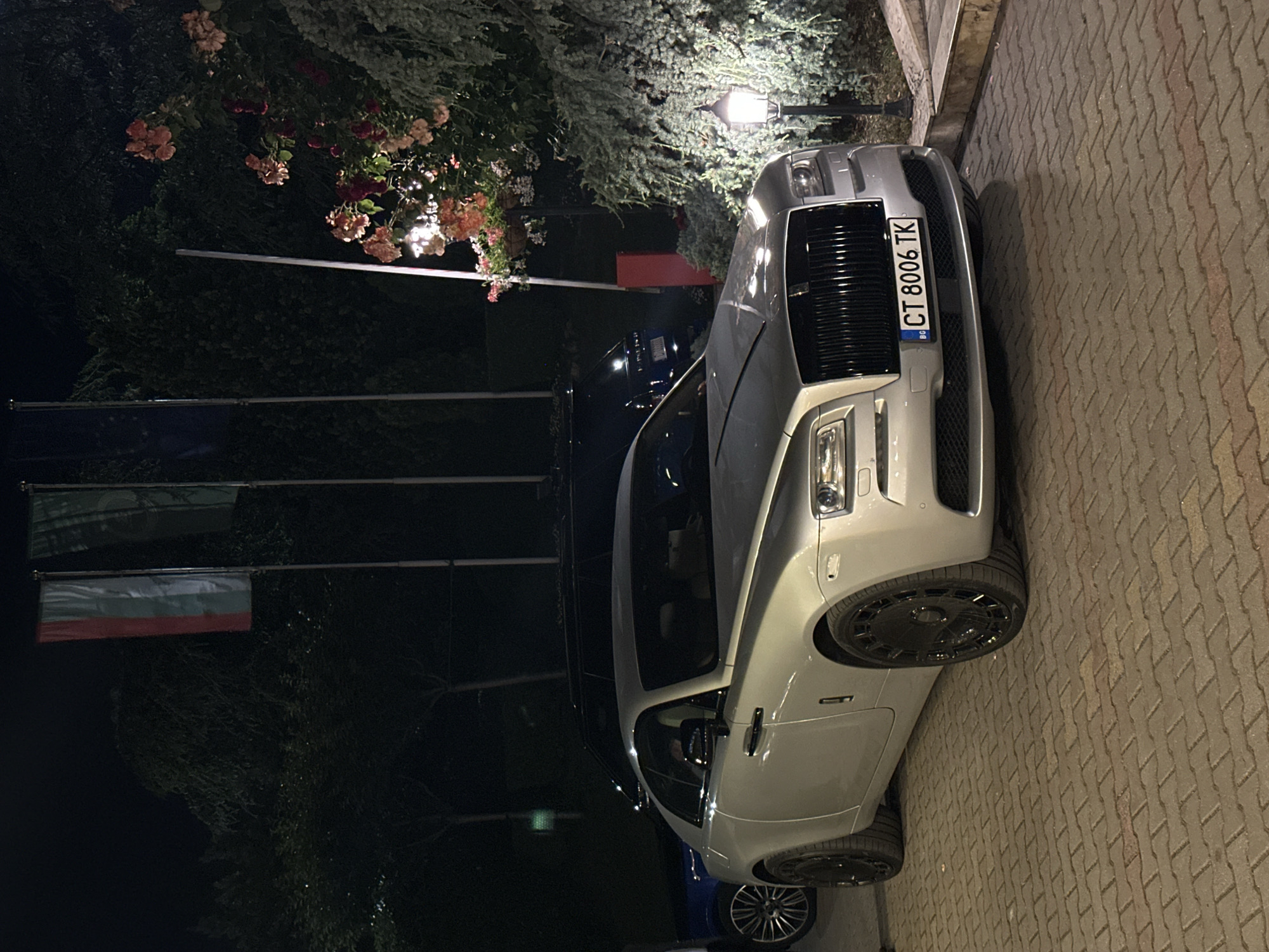 The image depicts a silver-colored Rolls-Royce Wraith coupe parked at night on a herringbone-patterned brick paver surface in Stara Zagora, Bulgaria. The vehicle's front license plate, "BG CT 8006 TK," identifies it as registered in Bulgaria, with "CT" indicating the Stara Zagora region. The Rolls-Royce is fitted with dark, multi-spoke wheels.
In the background, against a dark sky, three flagpoles are visible. The leftmost flag displays horizontal stripes of red, white, and green, consistent with the flag of Hungary. The middle flag is dark green, and the rightmost flag is dark blue, both without discernible details. Dense vegetation, including trees and bushes, forms the backdrop and extends to the right side of the frame. Artificial illumination highlights sections of the scene; a bright light source illuminates a cluster of red and pink roses on a bush on the right, and a decorative, dark-colored lamppost on the lower right casts light onto the adjacent greenery and pavement.
Partially visible behind the Rolls-Royce is a dark-colored SUV, and a wheel belonging to another vehicle is seen on the far left. A vertical red object, potentially part of a barrier system, is positioned in the mid-ground. No individuals are present in the image.