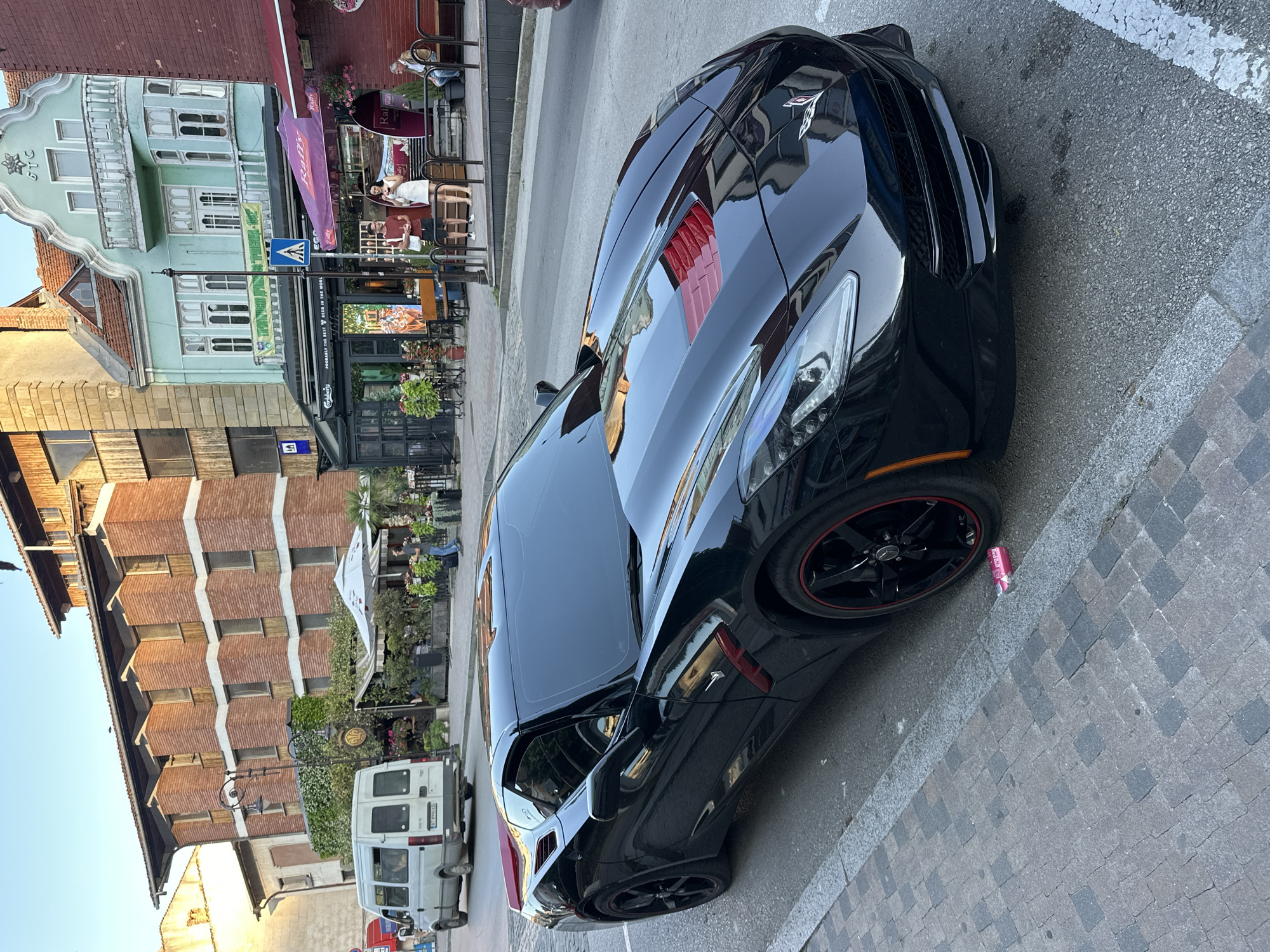 A black C7 Chevrolet Corvette, featuring red accents on the hood vents and red pinstripes on its black multi-spoke wheels, is parked parallel to a cobblestone curb on the right side of a paved street in Veliko Tarnovo, Bulgaria. The car's windows are tinted, and its front bumper is visible. A pink Red Bull can lies on the cobblestone sidewalk next to the front passenger wheel.
The street is lined with multi-story buildings. To the left of the frame, a white utility van with the Bulgarian license plate "BT 8990 CC" (confirming Veliko Tarnovo) travels away from the viewer. Behind the car, a tall building constructed of reddish-brown brick with prominent horizontal white bands and distinctive angled window structures on its upper floors is partially visible. The ground floor of this building features a restaurant or cafe with extensive outdoor seating, lush green foliage, and white umbrellas.
Further down the street, on the right side, stands a three-story mint green building with ornate architectural details, including a decorative balcony and arched windows. A Cyrillic banner above the ground floor reads "ТРИАТЛОН ДРУЖБА - 1982" (Triathlon Friendship - 1982), and "БТС" is visible near the roofline. The ground floor of this building houses a shop with a "Carlsberg" sign stating "PROBABLY THE BEST BEER IN THE WORLD" and another adjacent establishment under a purple "Rally" awning.
In front of these shops, near a pedestrian crossing sign, a man in a red shirt and shorts stands alongside a woman in a white dress who is holding a phone. Another man in a dark shirt and jeans walks on the sidewalk further down the street. An older man sits at an outdoor table under a red awning to the far right. The scene is illuminated by late afternoon or early evening sunlight, casting soft shadows.