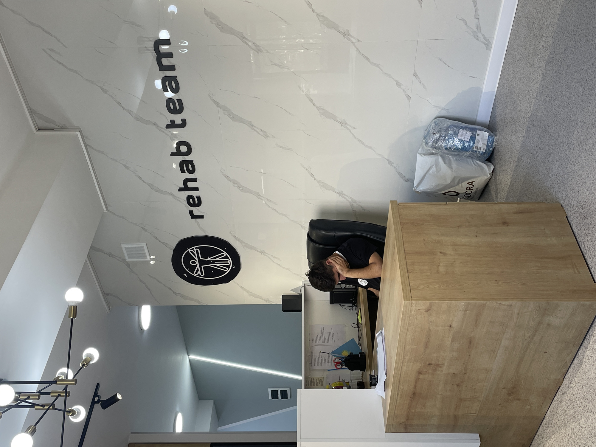 A young man in a black t-shirt is asleep at a wooden reception desk in an office. The man's head is resting on his hand, and his eyes are closed. The reception desk has papers, scissors, and a flag on it. In the background, the wall is covered in white marble tiles with gray veins, and a black sign with the words "rehab team" is mounted on the wall. To the left of the desk, there is a modern chandelier with multiple light bulbs. The office floor is carpeted with a gray speckled pattern. In the lower right corner, two large white bags are leaning against the wall. The overall scene suggests a quiet, possibly late-night or early-morning shift at a rehabilitation center or medical office. The specific location in Almaty, Kazakhstan, cannot be determined from the visual information provided in the image.