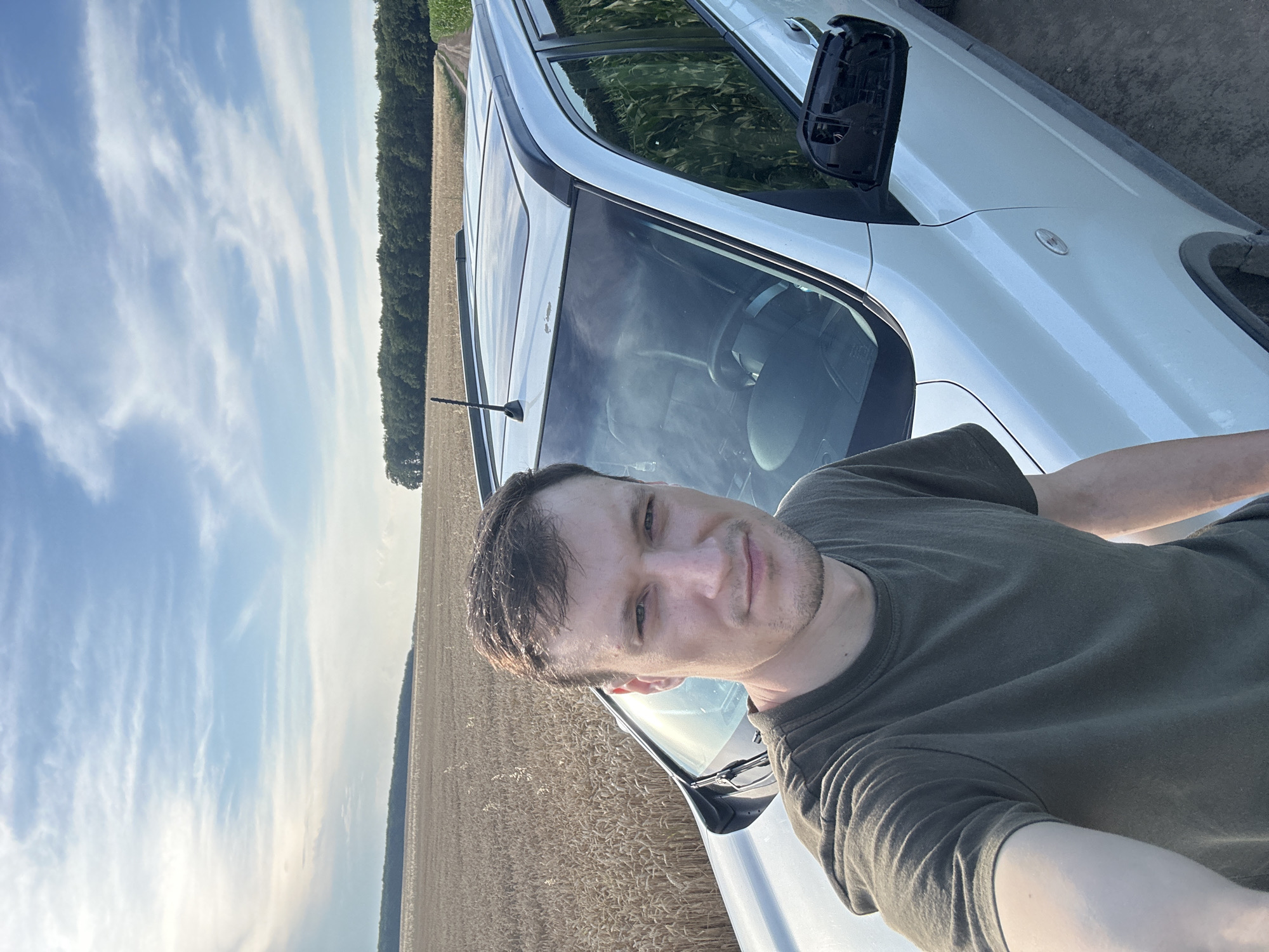 In the city of Makiv, Ukraine, a young man with short brown hair and a light complexion is captured in a close-up selfie. He is wearing a dark green t-shirt and is positioned in front of a white SUV. The car's hood and windshield are visible, reflecting the bright sky. Behind the car, a vast field of wheat stretches towards a distant treeline. The sky above is a mix of blue and white, with wispy clouds. The overall scene is one of rural landscape during what appears to be daytime.