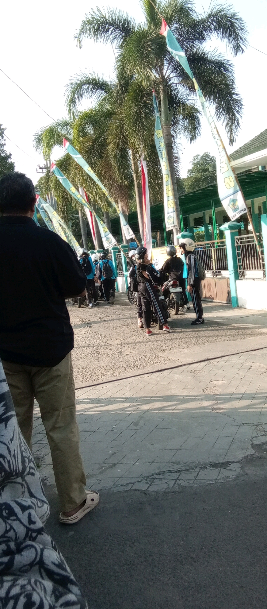 The image depicts an outdoor scene in Kediri City, Indonesia, likely in front of a school or public building, suggested by the green fence and building visible in the background. A group of young people, dressed in dark athletic wear and helmets, are gathered around motorcycles. Some are interacting with each other, while others appear to be preparing to depart.
In the foreground, a person in a black shirt and khaki pants is facing away from the camera, observing the group. The environment features a paved walkway with a textured surface, and a darker asphalt area. Tall palm trees with lush fronds line the background, interspersed with numerous colorful flags that appear to be celebratory or promotional, featuring text and designs. The overall lighting suggests it is daytime, with sunlight casting shadows. The presence of motorcycles and the attire of the younger individuals imply a context related to transportation, possibly for commuting to or from an educational institution.