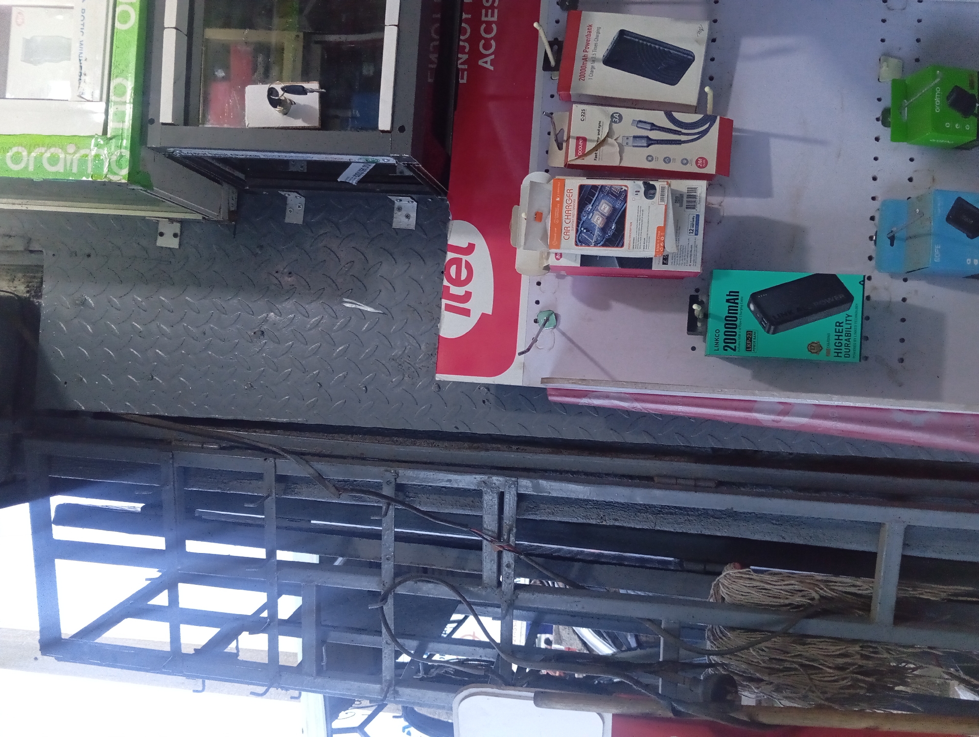 The image depicts an indoor environment, likely a shop, with a focus on electronic accessories. In the foreground, a metal shelving unit is visible, with various items displayed on it. To the left, a mop with a frayed head hangs from a hook. Mounted on the wall is a display of packaged electronic goods, including power banks and car chargers. Brands like "itel", "LINKCO", and "oraimo" are discernible. The background shows a grey metal wall with a diamond plate texture and what appears to be a ladder or scaffolding structure. The lighting suggests an indoor setting, possibly with some natural light filtering in. There are no people visible in the image. The overall scene suggests a retail space specializing in mobile accessories.