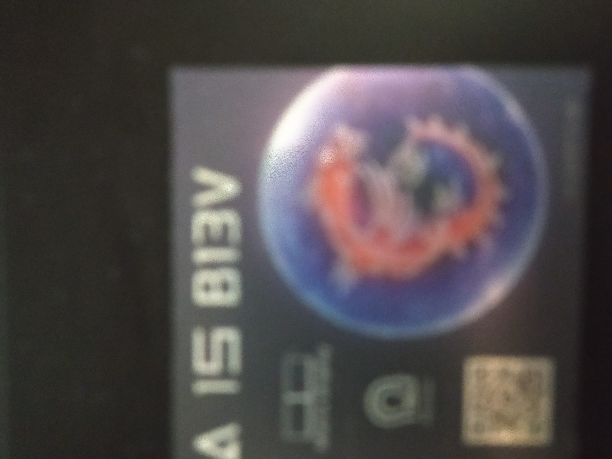 The image shows a blurred sticker. The sticker has the text "AI 15 B18V" vertically on the left side. The right side of the sticker features a round image with a blue background and an orange shape inside, possibly resembling a dragon or creature. Below the image are two indistinct logos or symbols and a QR code in the bottom right corner. The location of the image is unknown; there is no information to indicate it's in Sofia, Bulgaria.