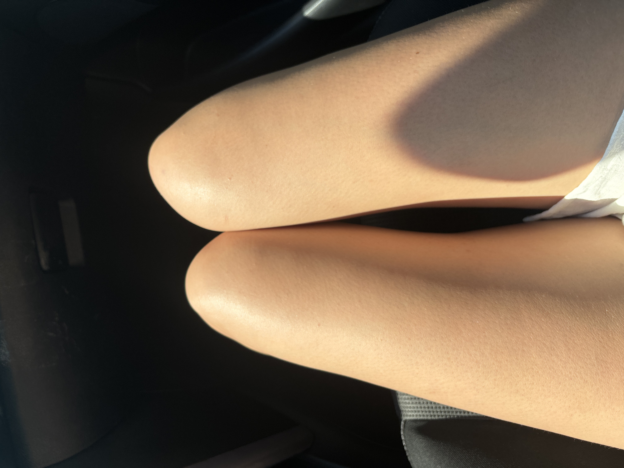 The image shows a close-up of a person's bare legs, with the skin illuminated by sunlight casting a shadow across the upper thigh. The setting appears to be indoors, possibly a car interior, indicated by the dark, textured surface on the left. The person is sitting, with their knees bent. A piece of white fabric is visible on the right side of the frame, possibly clothing or a seat cover. The skin texture is visible, with small bumps, and there is a slight sheen from the light. No text is visible in the image. The scene suggests a casual moment, possibly during daytime given the strong sunlight.