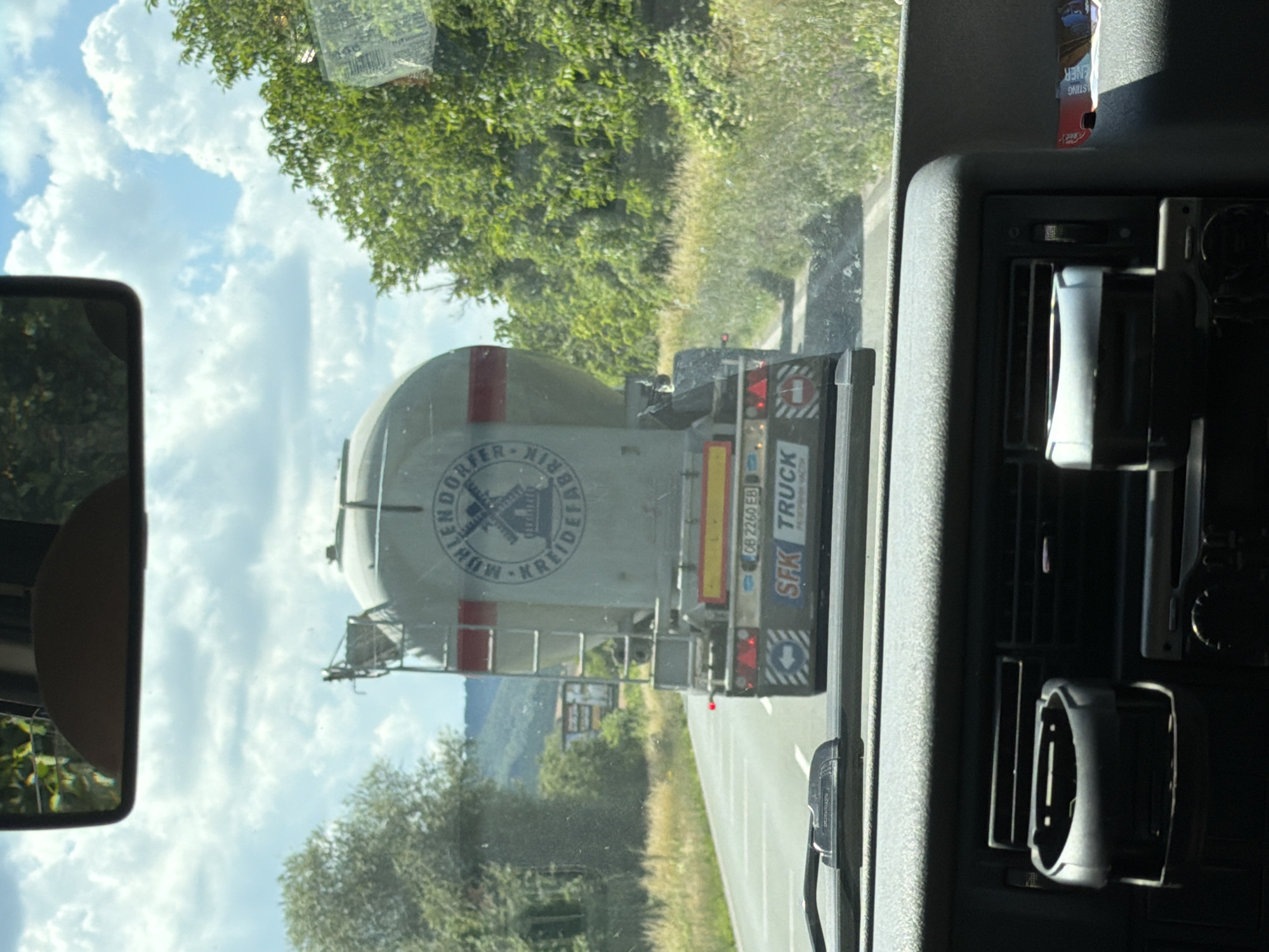 The image is taken from the interior of a vehicle, looking forward through the windshield and the interior dashboard. The vehicle is on a road, likely in a rural area. In front, a white cement mixer truck is visible, driving on the same road. The truck has a circular logo on its side with a windmill design and text that appears to read "MUHLENDORF KREIDEFABRIK". The license plate of the truck is visible, reading "OB 2260 EB". The truck also has "SFK TRUCK" written on its rear. The environment outside the vehicle consists of green trees and vegetation lining the sides of the road, with a cloudy blue sky above. The dashboard of the vehicle in the foreground shows a cup holder and air vents. The location is identified as Riahovtsite, Bulgaria.