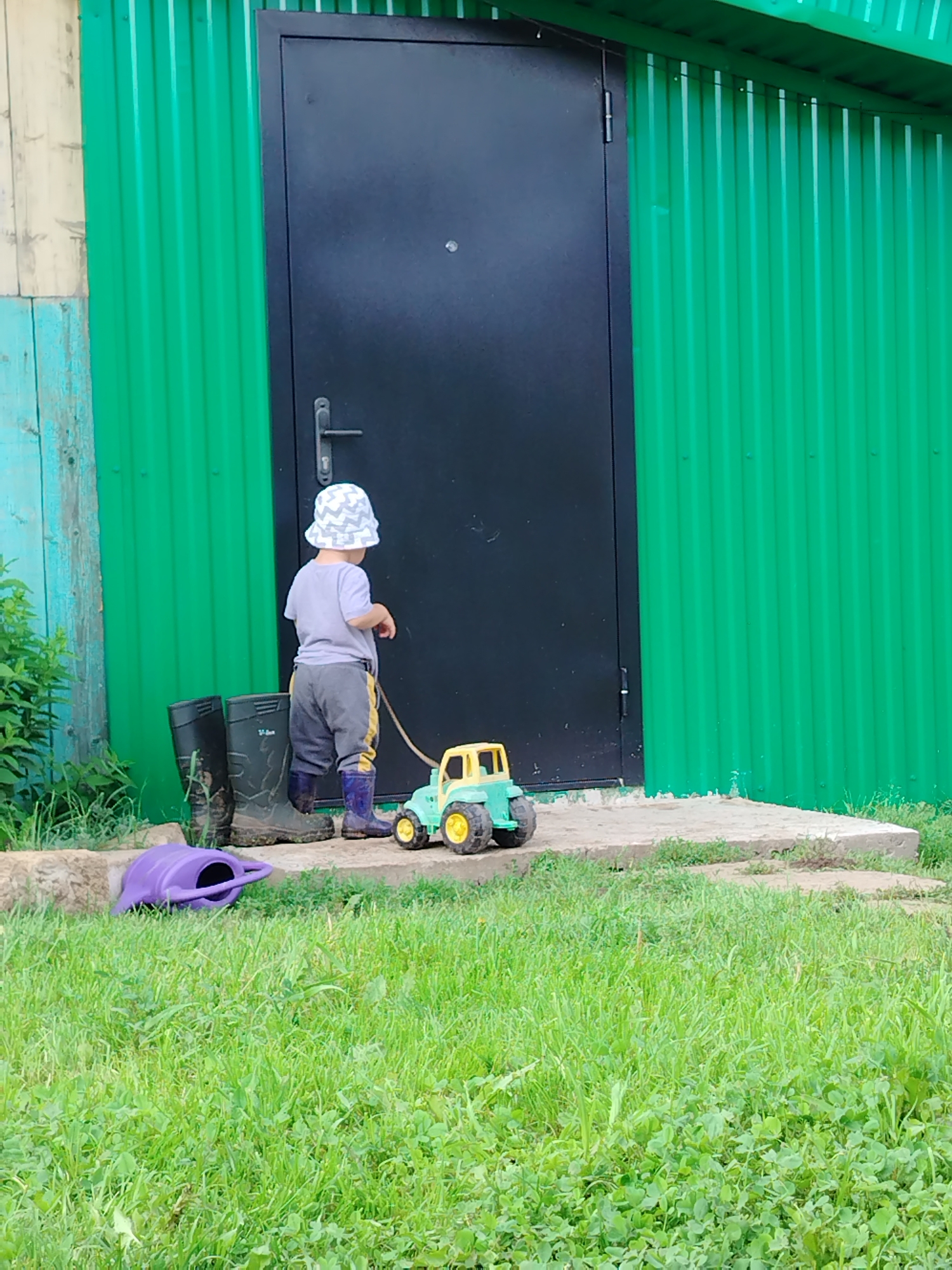 The image depicts an outdoor scene, likely in or around the village of Gozhan, Russia, on a bright day. The foreground is dominated by lush green grass with some clover. A small, brightly colored toy tractor, predominantly yellow and green, rests on a concrete path. To the left of the path, a purple watering can is partially visible in the grass. Further back, on the concrete path, stand two pairs of adult-sized black rubber boots and one pair of blue children's boots.
Behind the boots, a young child, wearing a grey t-shirt, grey pants, a patterned bucket hat, and blue boots, stands facing away from the camera. The child appears to be interacting with the toy tractor, with a string or rope extending from the child's hand to the tractor. Behind the child and the path is a large, dark metal door, set into a bright green corrugated metal structure. To the left of the door, a section of a wooden wall with visible planks, painted in alternating light and teal colors, is present. The overall impression is of a child playing outdoors near a building, with gardening tools and toys nearby.