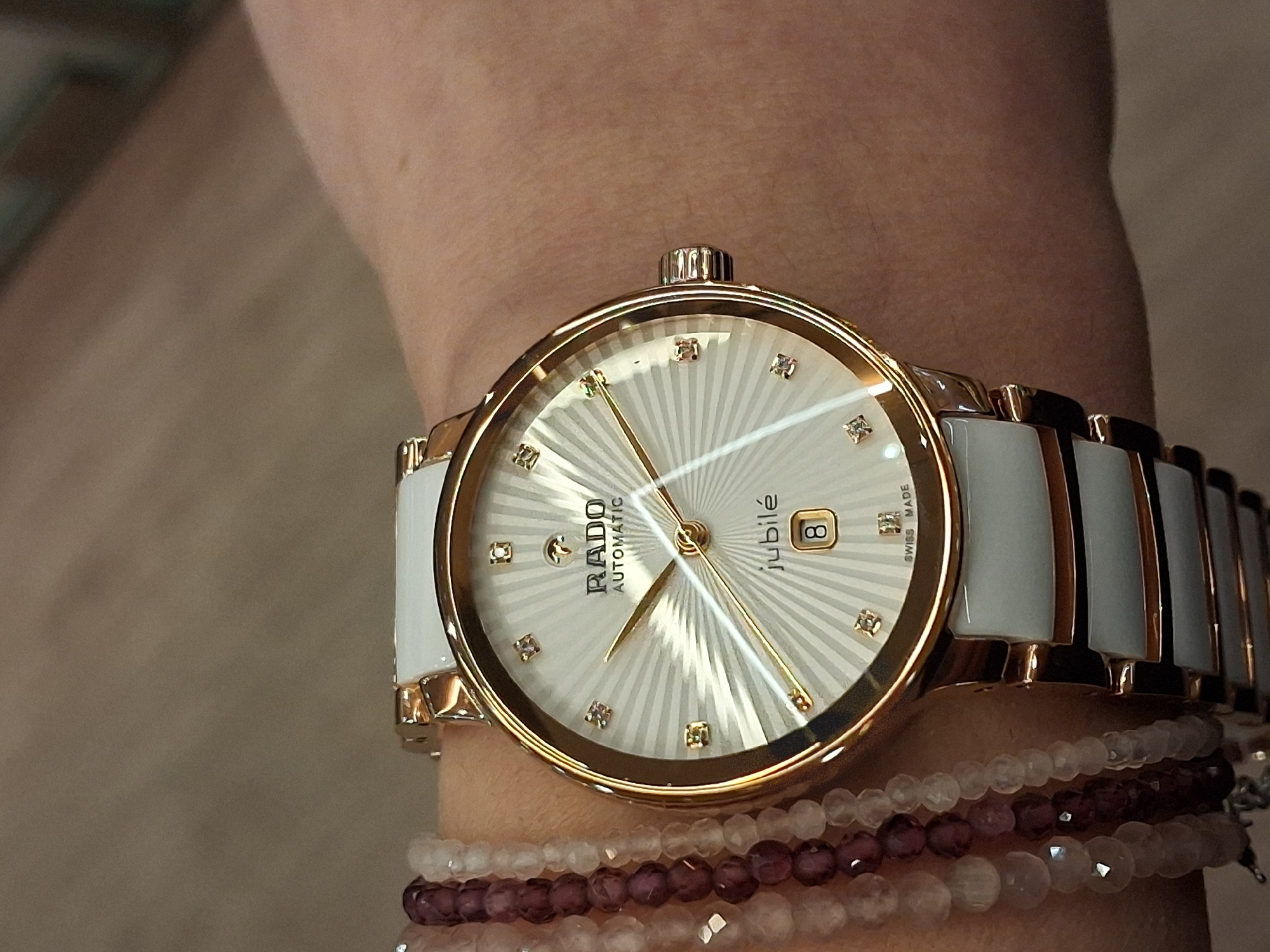 A human left wrist, exhibiting fair to light brown skin tone, is centered in the frame. A Rado Jubilé Automatic wristwatch is worn on the wrist. The watch features a circular gold-toned case and a fluted gold-toned crown on the right side. Its white dial displays a radial sunray pattern with "RADO AUTOMATIC" inscribed at the 12 o'clock position and "jubilé" above a date window showing the numeral "8" at the 6 o'clock position. "SWISS MADE" is visible at the bottom of the dial. Eleven square-cut diamonds function as hour markers. The watch has gold-toned hour, minute, and second hands. The bracelet consists of alternating white ceramic segments and gold-toned metal links. To the left of the watch, three beaded bracelets are worn: one with small, dark reddish-brown, faceted beads, and two with pale pink to clear, faceted spherical beads. The background is uniformly blurred, appearing as an indistinct light brown or beige surface, consistent with an indoor environment. No specific visual information within the image confirms the location as Varna, Bulgaria.