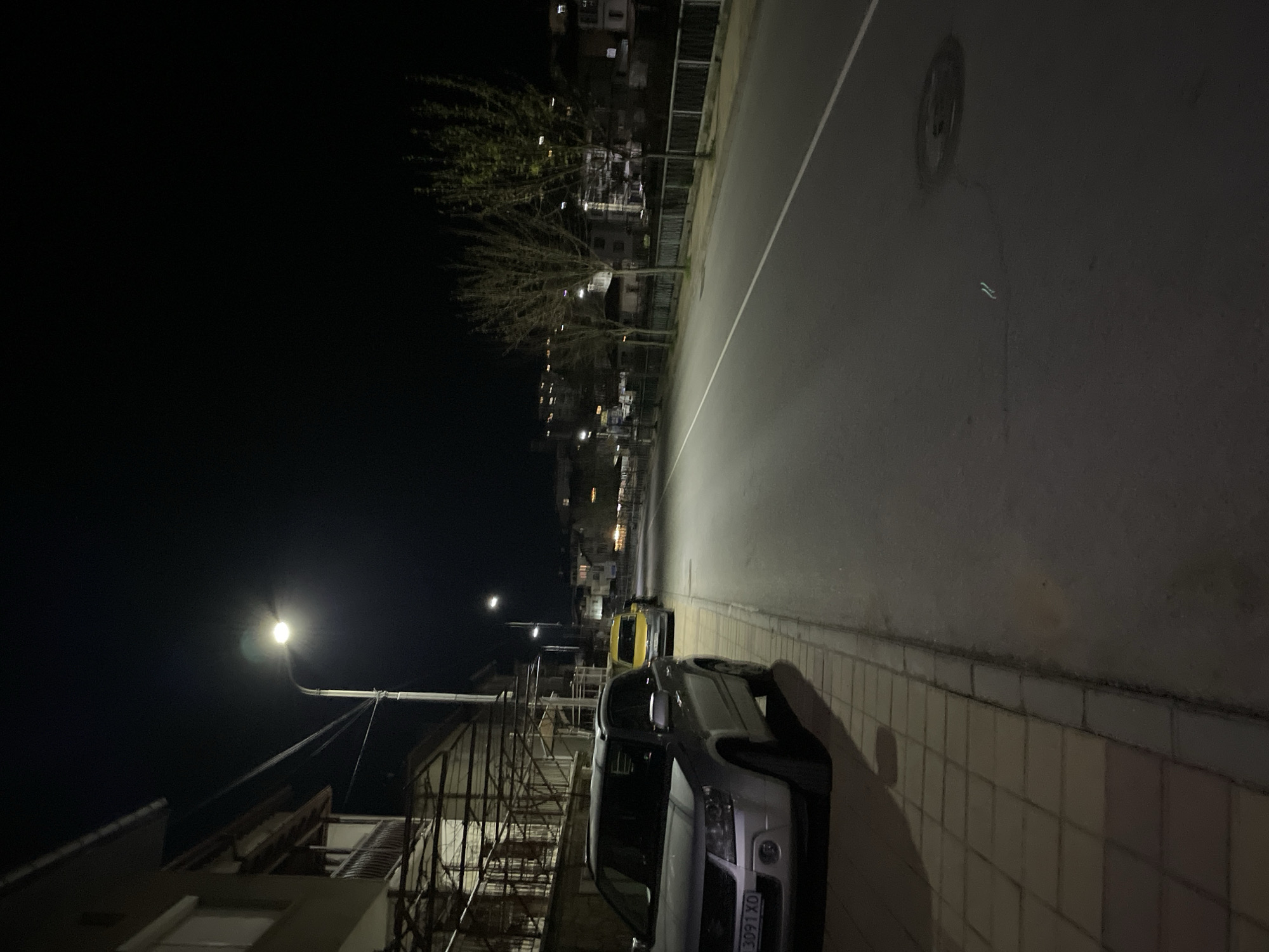 The image depicts a night scene in Zlatograd, Bulgaria. A street is lined with buildings, with a parked silver car and a yellow vehicle visible on the right side. Streetlights illuminate the scene, and bare trees line the road further in the distance. The location seems to be a residential or commercial area based on the presence of buildings with visible windows.