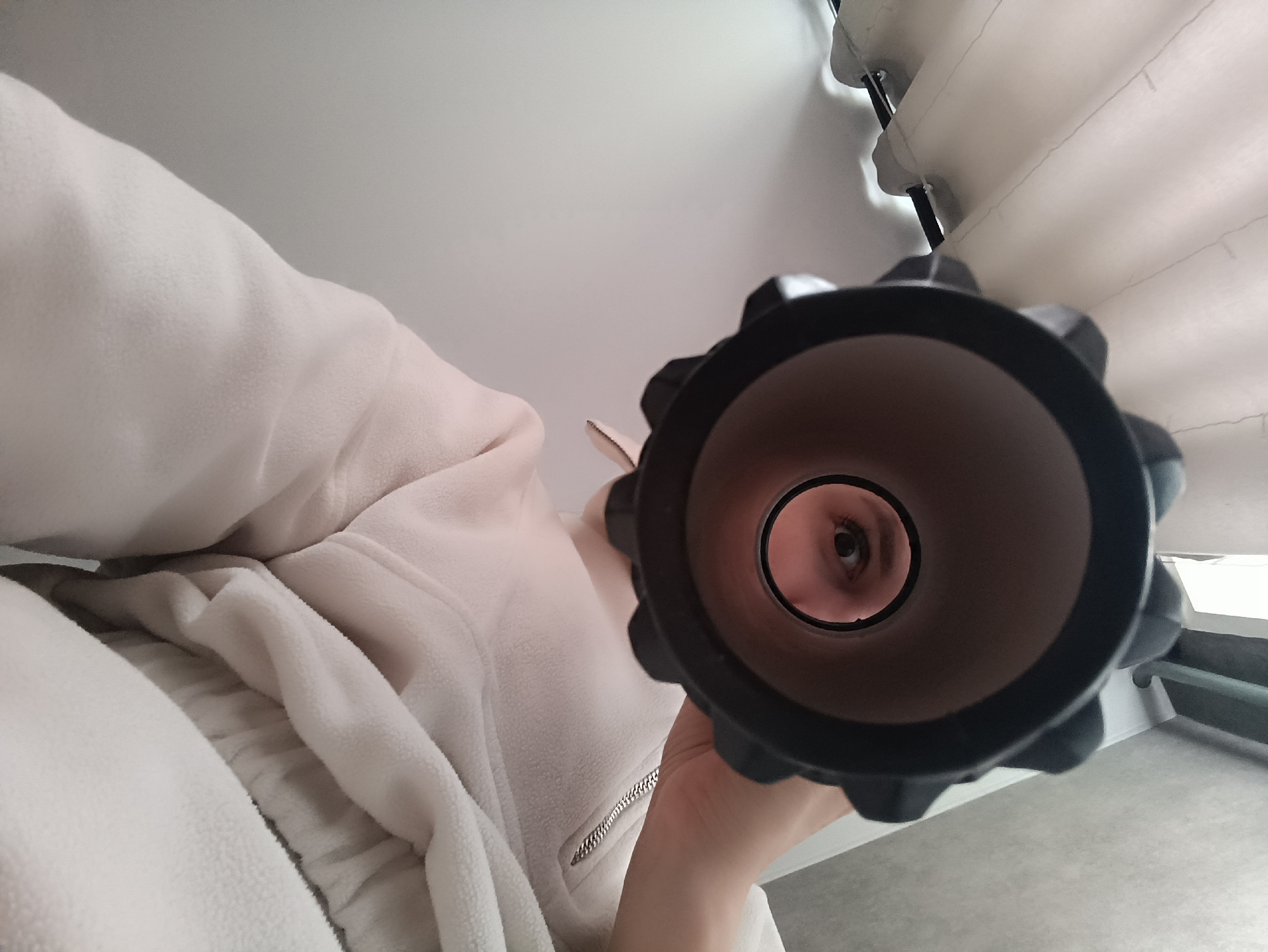 The image captures a low-angle, close-up perspective of a person looking directly through a dark cylindrical object. The person, likely wearing a light-colored, possibly fleece, zippered jacket with a silver zipper, holds the cylinder in their hand. Only one of their eyes is clearly visible through the opening of the cylinder, looking towards the viewer. The object appears to be black, possibly a foam roller or a similar thick-walled tube, with a textured or ridged exterior.
The background suggests an indoor setting with light-colored walls and ceiling. In the upper right quadrant, white, layered, fabric-like structures are visible, possibly parts of blinds, curtains, or a light fixture, supported by thin black rods. The lower right corner reveals a light gray floor and a white baseboard, hinting at a room interior, possibly with a window nearby given the bright, diffused lighting. The overall lighting is soft and even, without harsh shadows, suggesting ambient light. The scene implies an experimental or playful act of looking through an object, offering a unique, tunnel-like view centered on the eye. There is no visible text in the image.